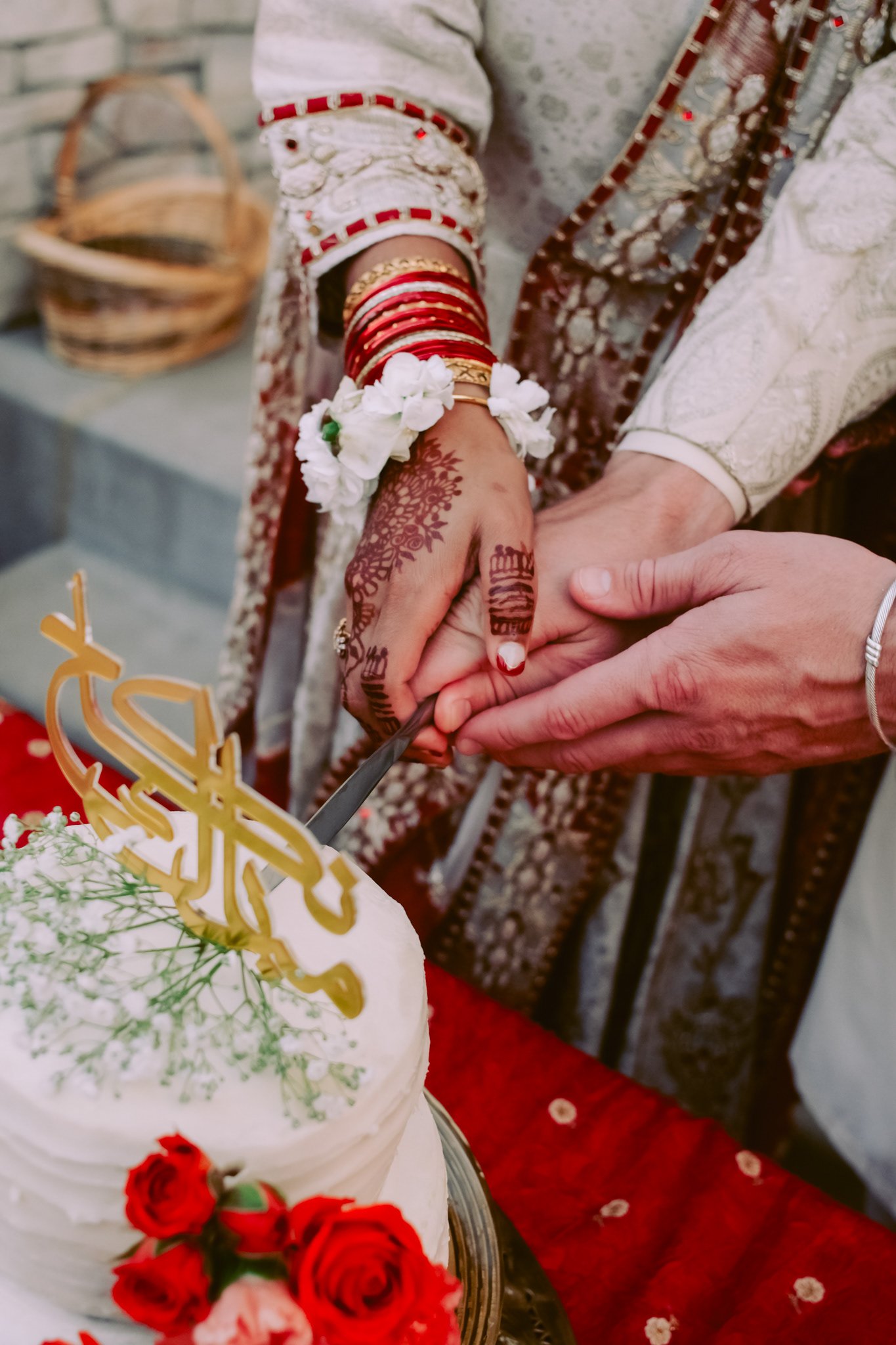 A bride and groom cut a wedding cake together, holding a cake knife. The bride has traditional henna on her hands, wears red bangles, and white floral bracelets. The groom wears a white patterned outfit. The cake is decorated with red roses and a gol