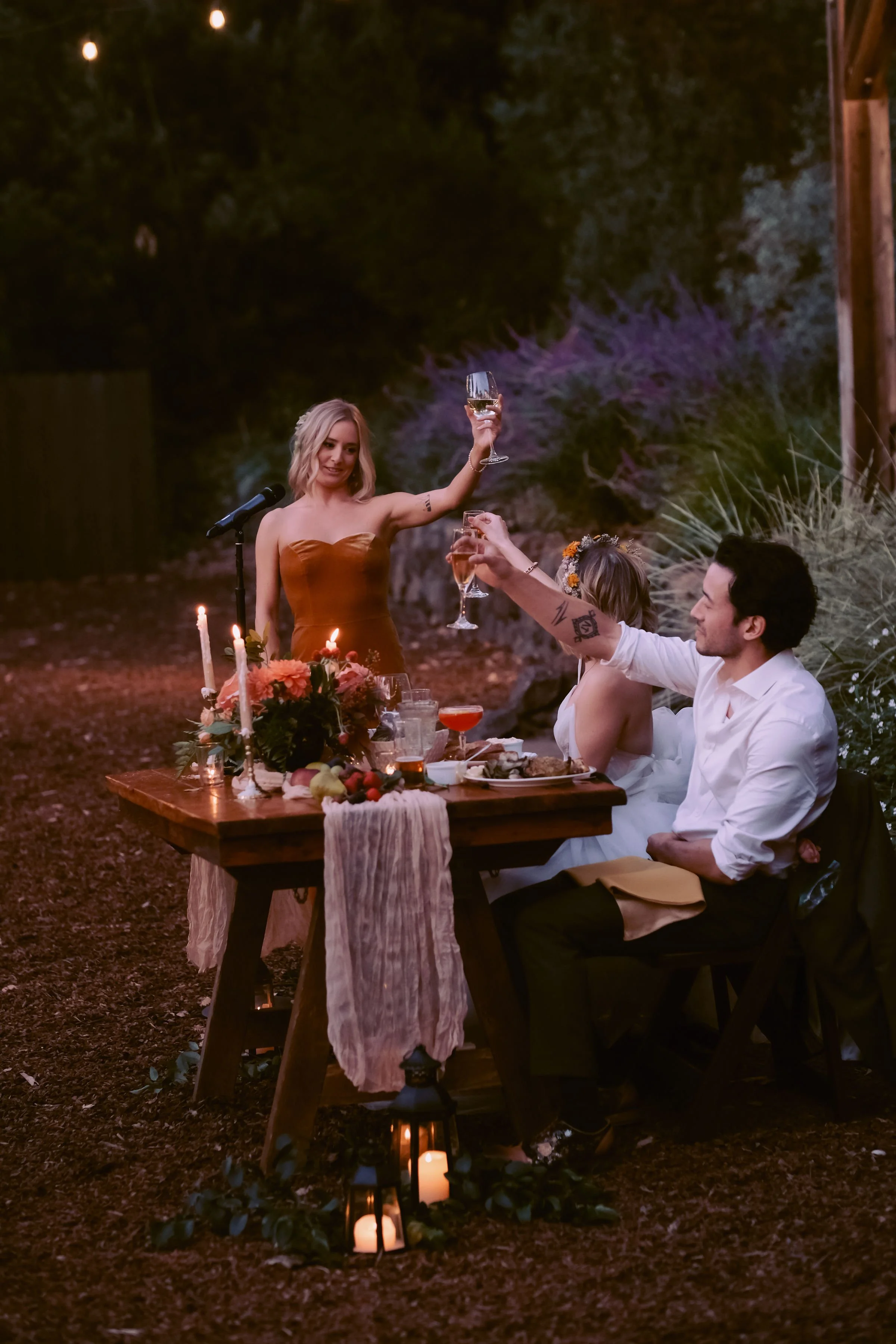 A group of four people celebrating at an outdoor dinner party during the evening, raising glass of wine for a toast with a decorated table with flowers, candles, and food.