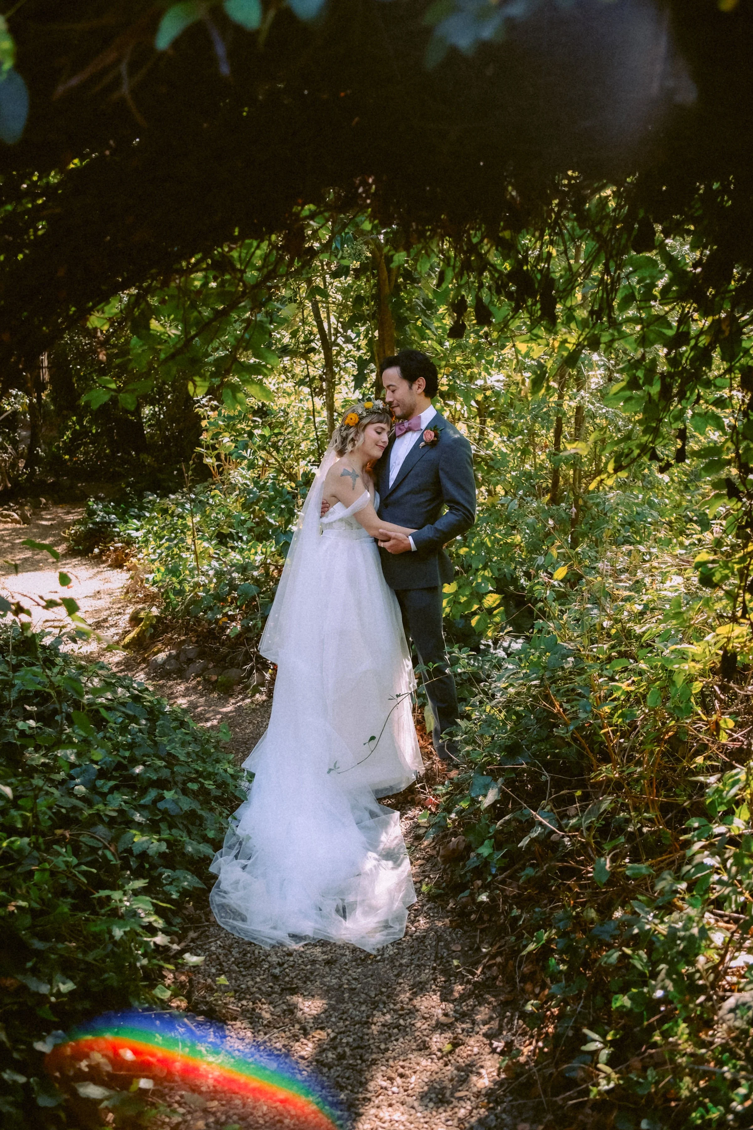 A bride and groom standing close together in a forest, surrounded by green foliage and sunlight, during their wedding photoshoot.