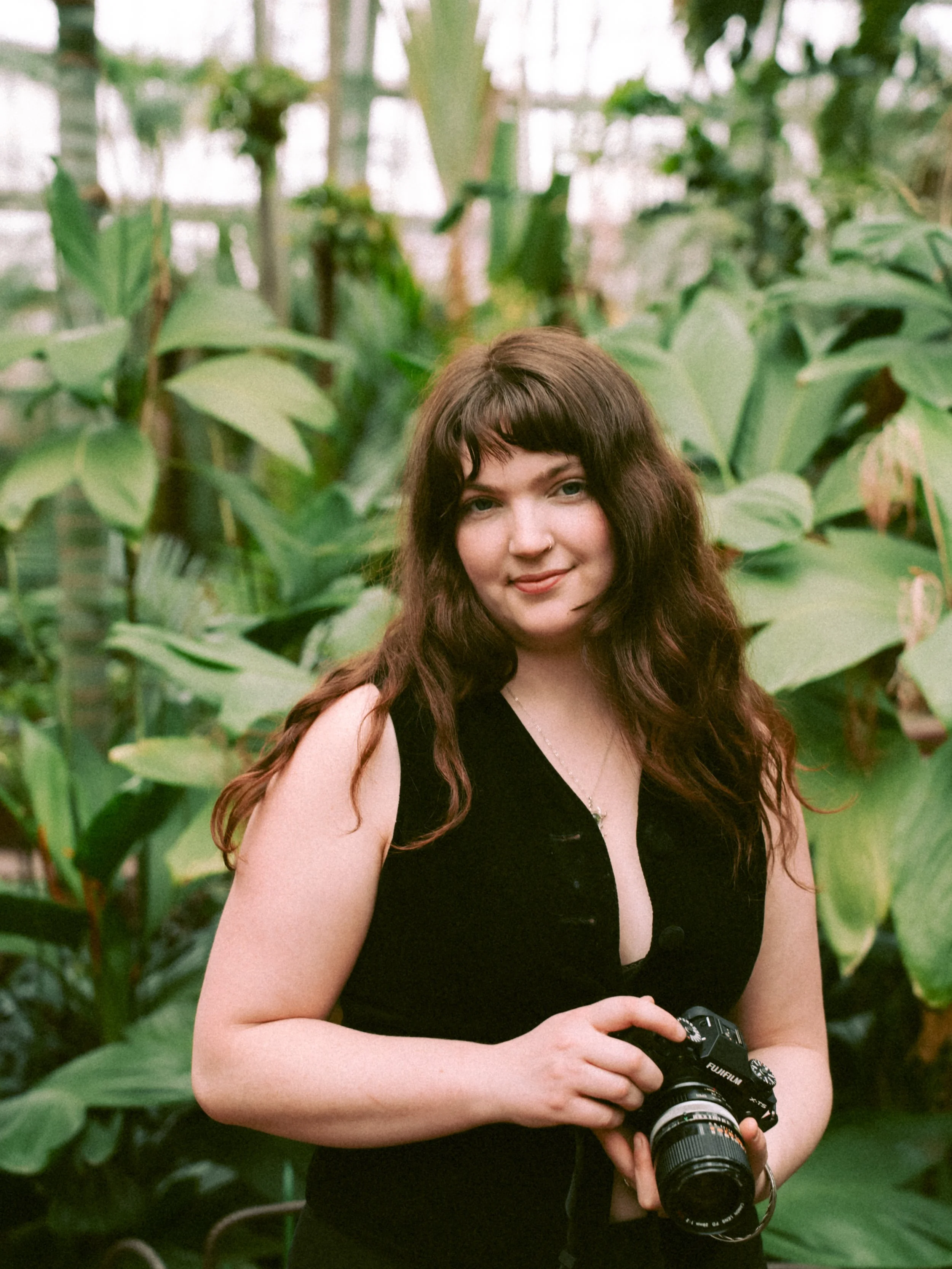 A young woman with long wavy brown hair wearing a black sleeveless top, holding a camera, standing in front of large green leaves of tropical plants.