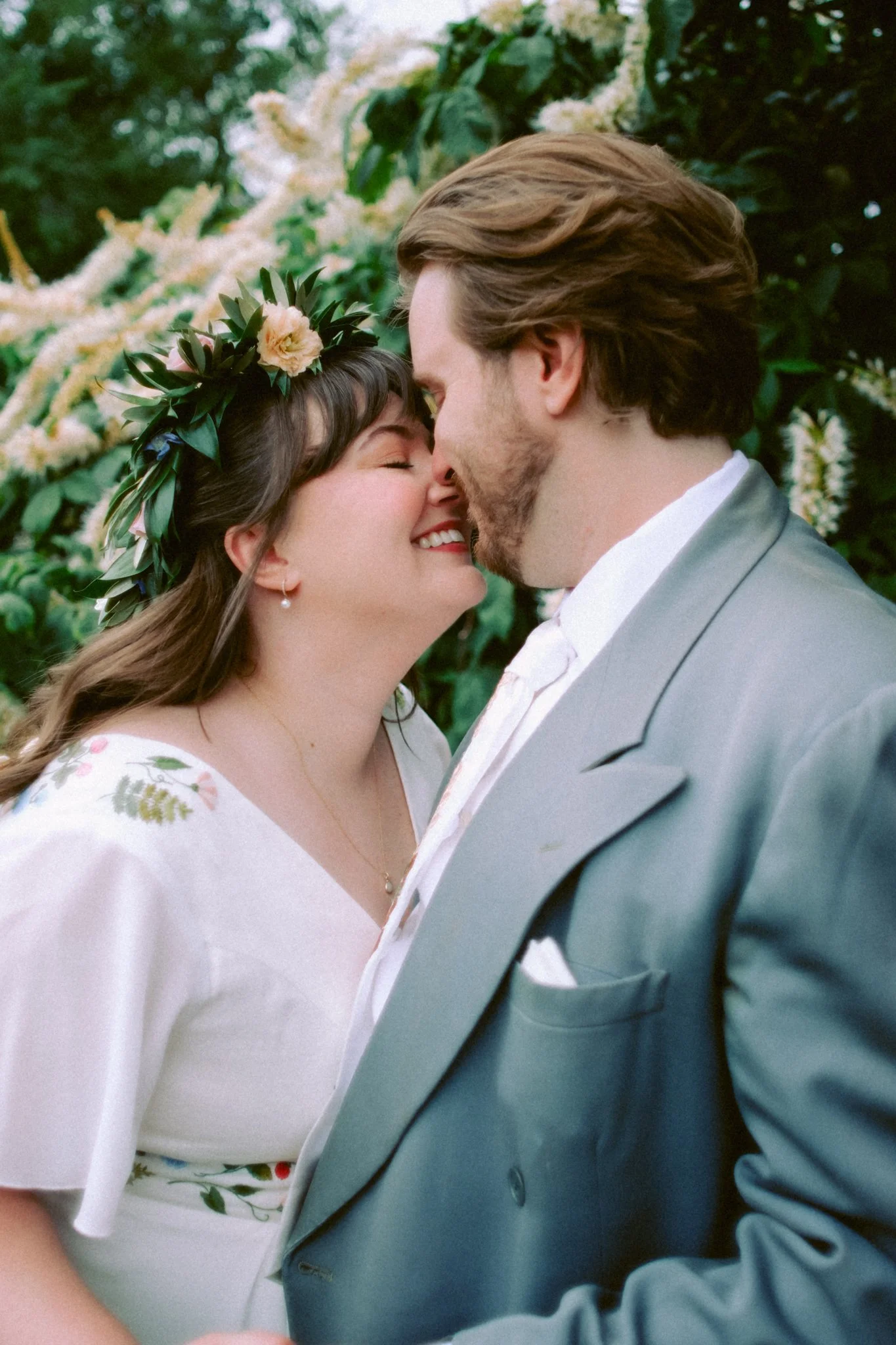 A couple shares a romantic moment with foreheads touching and eyes closed. The woman wears a floral crown and white dress, while the man is dressed in a gray suit. They are outdoors in front of lush greenery.