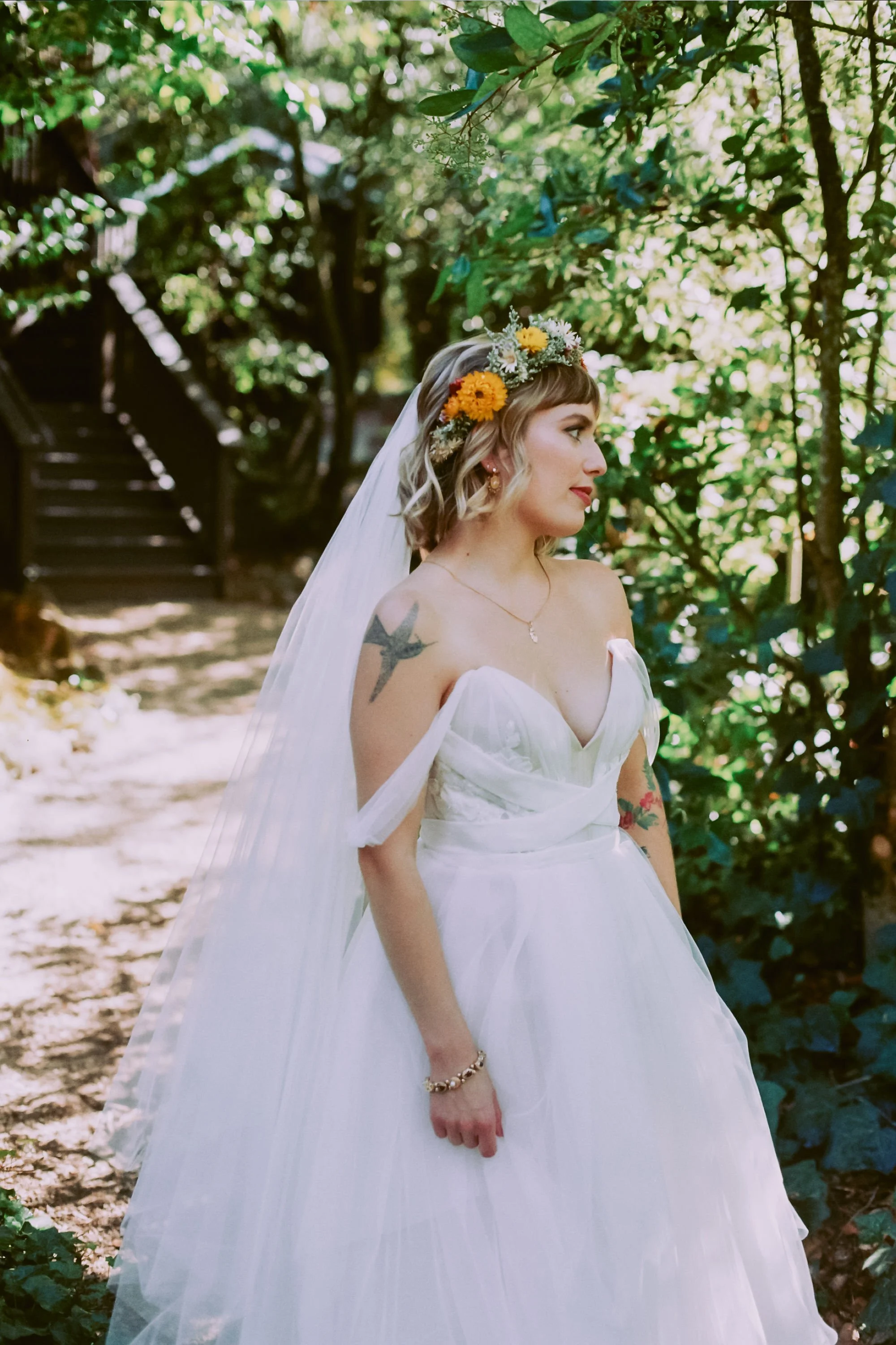 A woman dressed in a wedding gown with a floral crown, standing outdoors in a forested area with green leaves and sunlight filtering through.