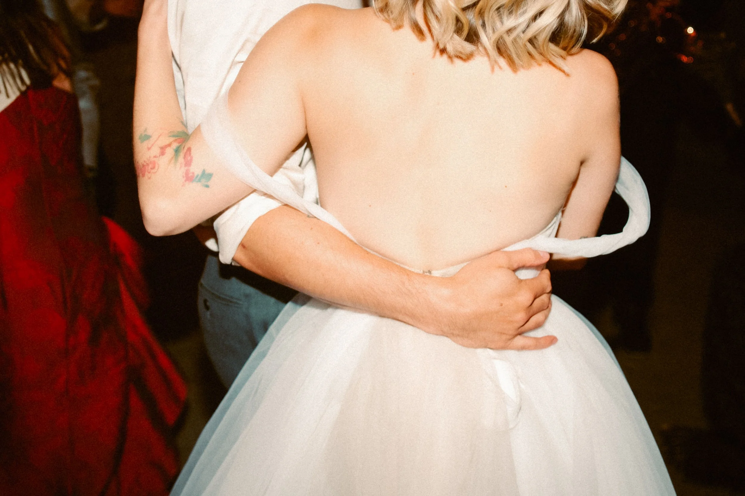 A couple dancing closely at a wedding reception, with the bride wearing a strapless white wedding gown and the groom in a dark suit. The groom's arms are wrapped around the bride's waist, and the bride's back is visible.