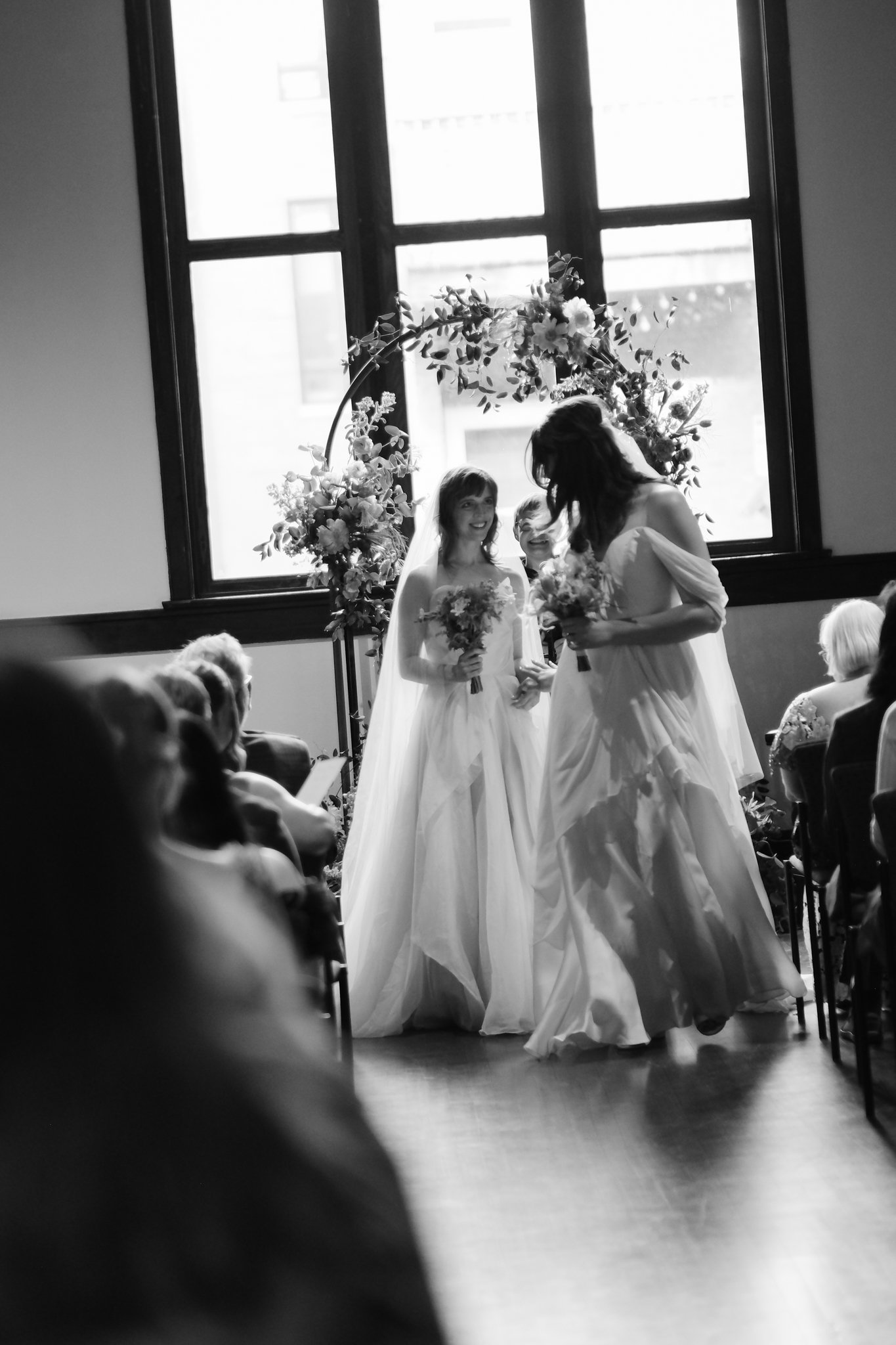 Black and white photo of a wedding ceremony with two brides exchanging vows, standing under a floral arch, with seated guests in the foreground.