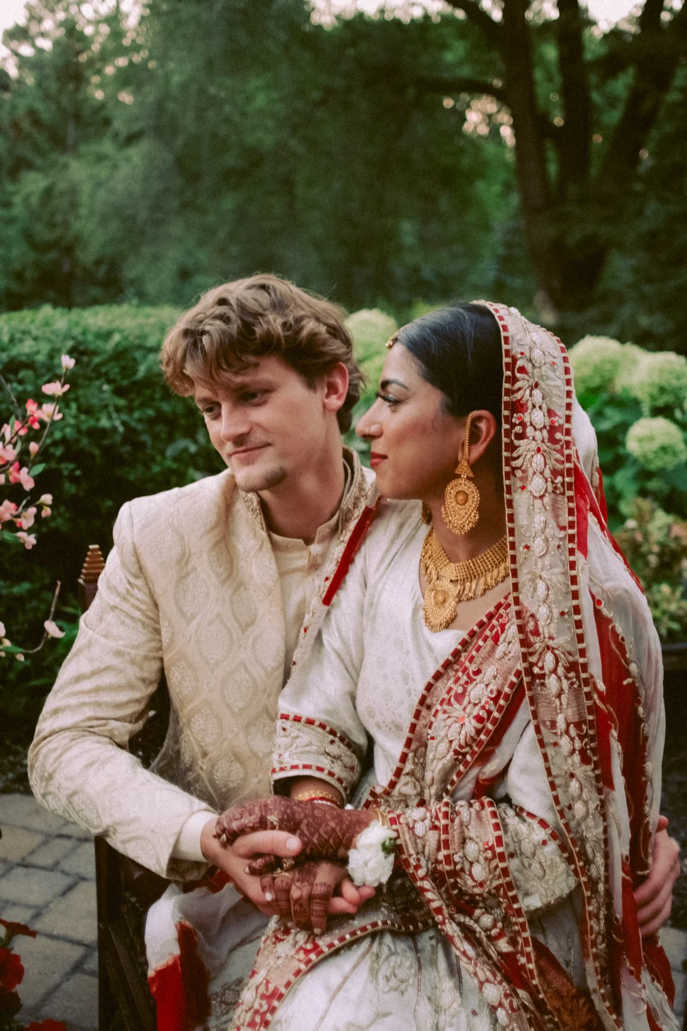 A man and a woman in traditional Indian wedding attire sitting outdoors, holding hands, with greenery in the background.