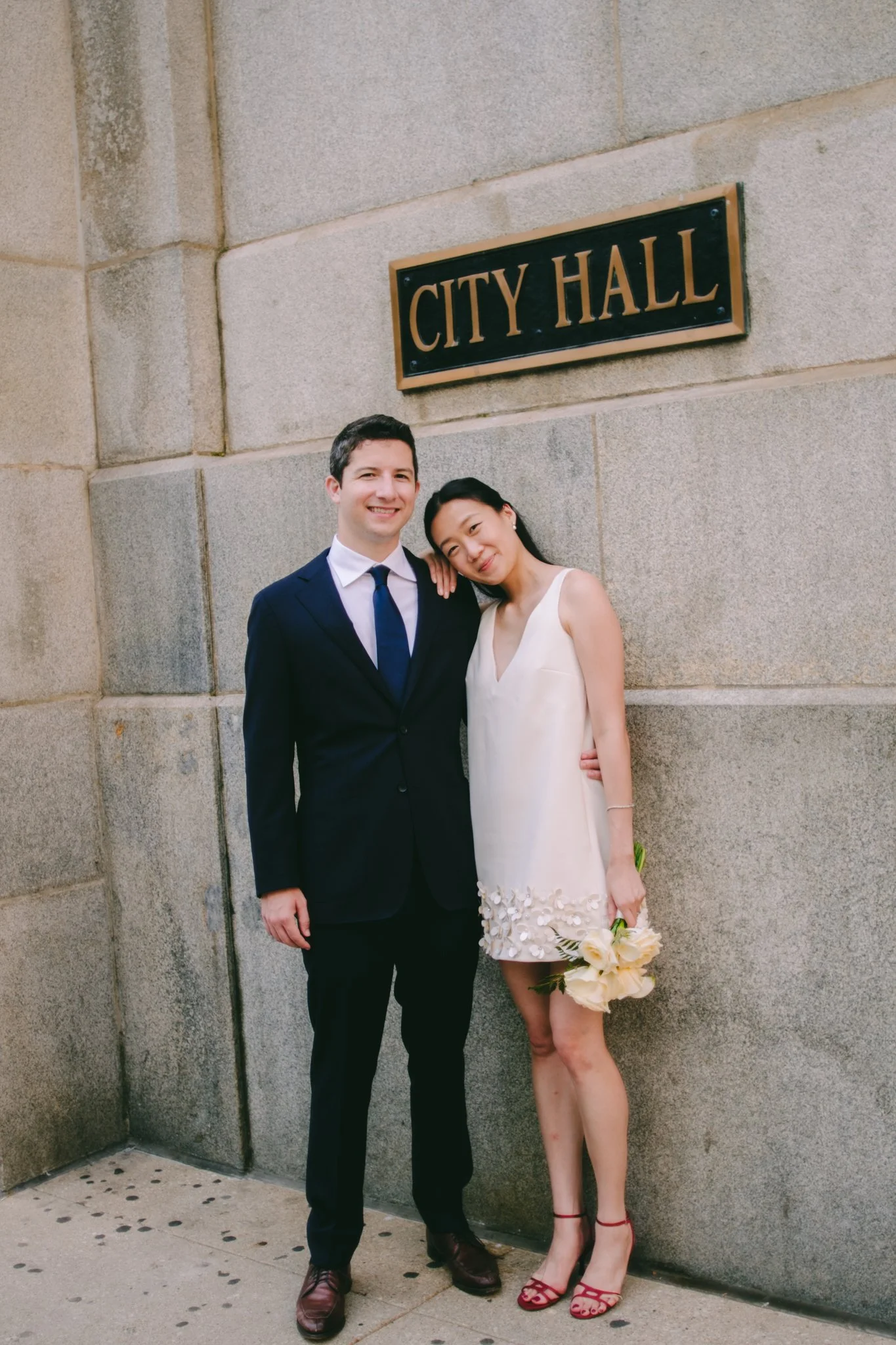 A smiling man in a dark suit and tie stands next to a woman in a cream dress holding a bouquet of white flowers, both leaning against a stone wall with a "City Hall" sign above them.