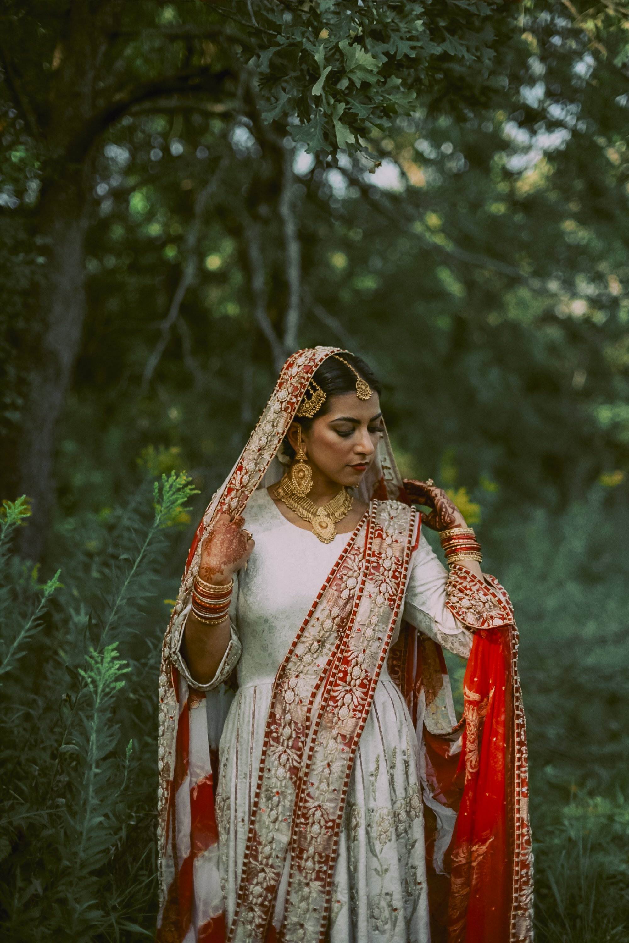 A woman dressed in traditional Indian attire, with intricate gold jewelry, a cream and red embroidered dress, and a matching head covering, standing outdoors among greenery and trees.