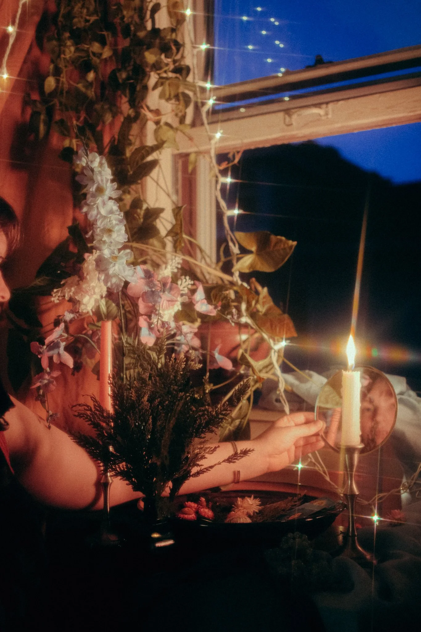 A person sitting at a table decorated with flowers, greenery, and a lit candle in a mirror holder, with string lights and an open window showing a dark blue sky outside.