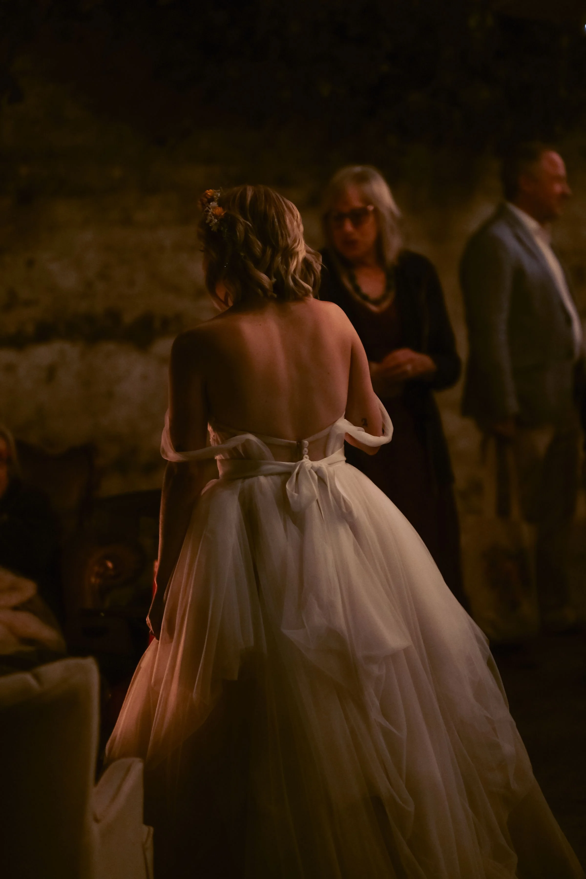 A woman in a strapless wedding dress with a full tulle skirt is seen from behind in a dimly lit room, possibly during a wedding or special event.