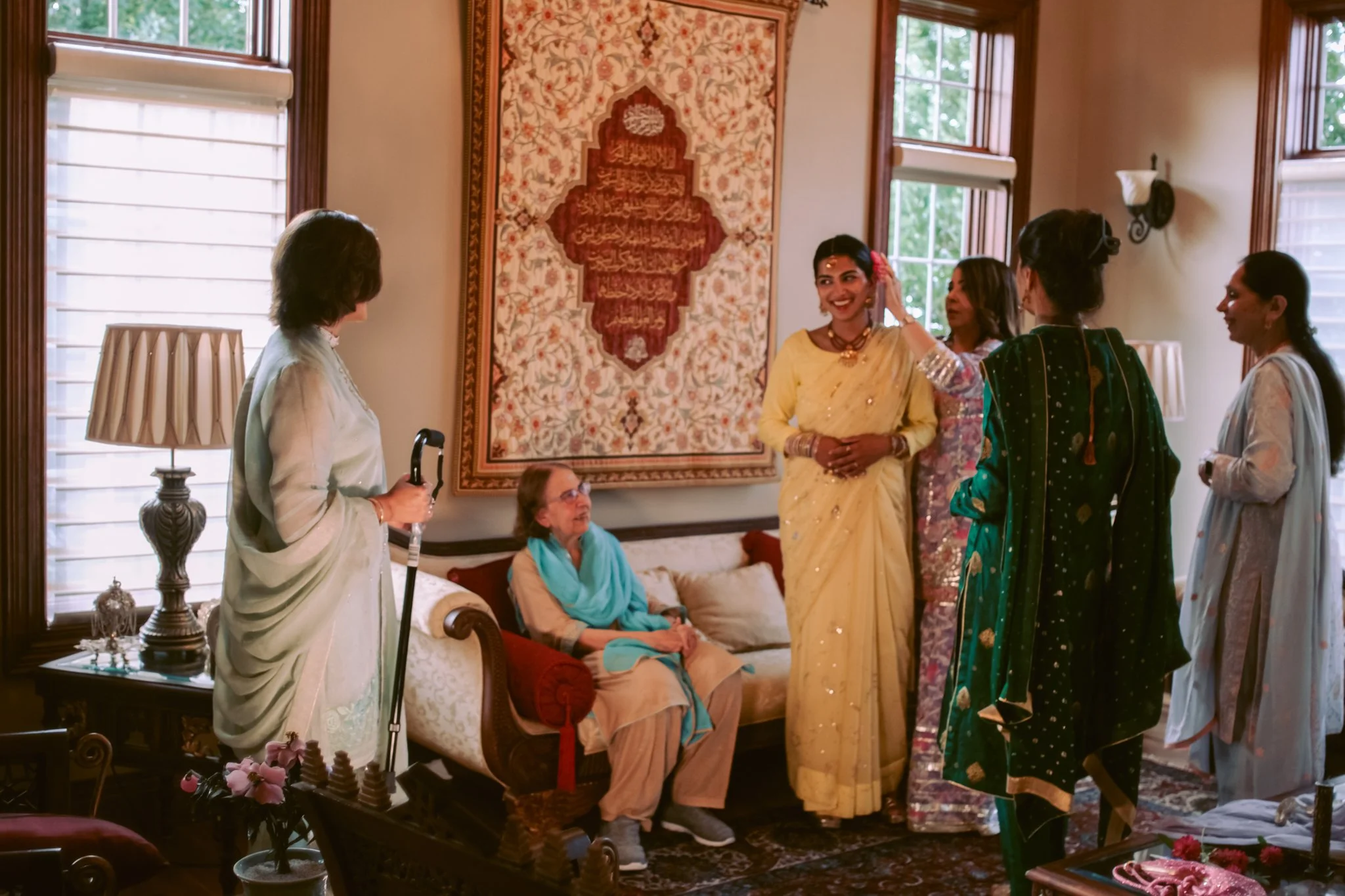 A group of women gather in a living room, with two women seated on a sofa and five women standing around, one of whom is wearing a yellow sari and smiling. The room has large windows, a patterned wall hanging, and traditional furniture and decor.