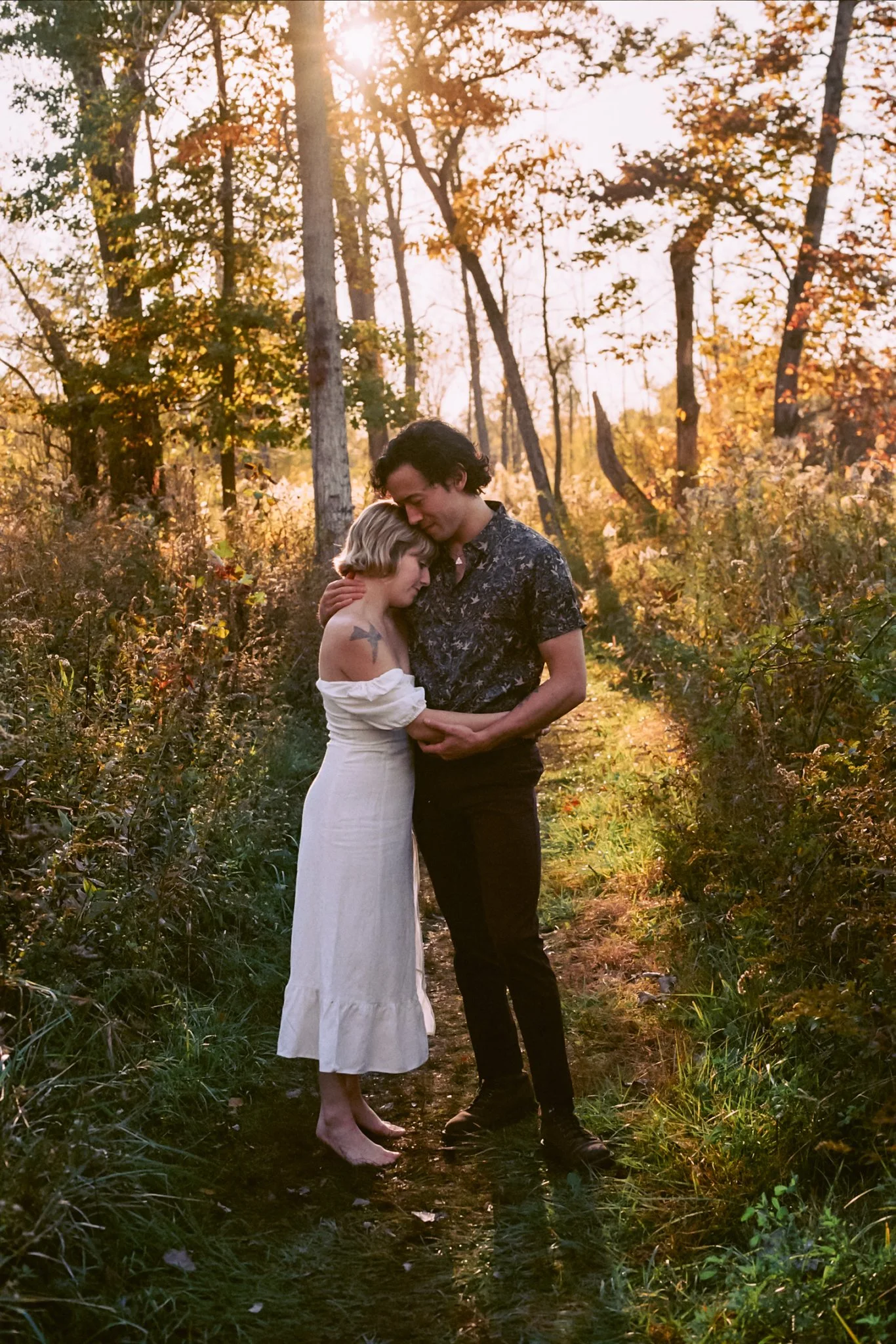 A couple embraces on a wooded trail during sunset, surrounded by autumn foliage.