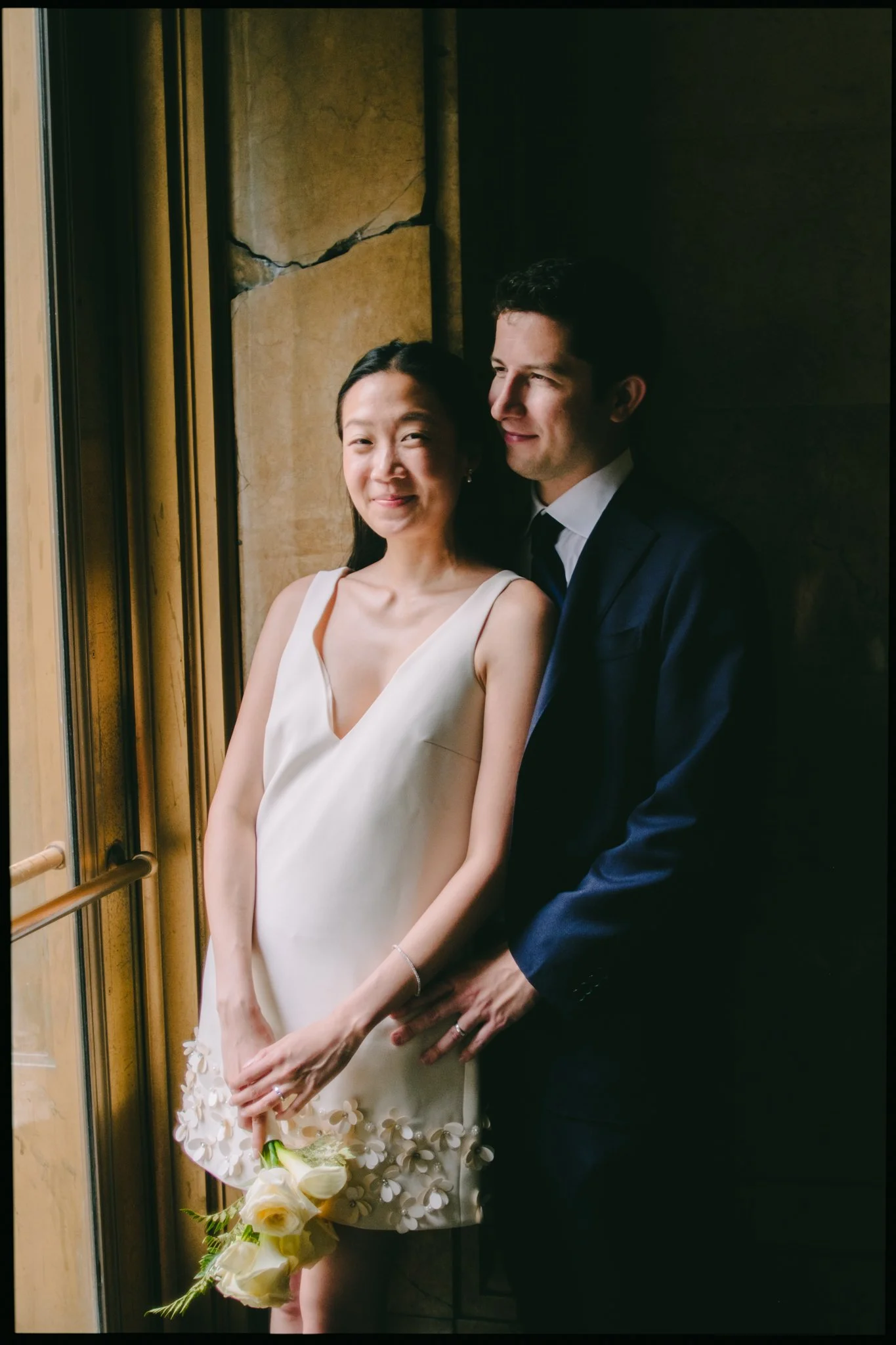 A joyful couple standing together, the woman holding a bouquet of white flowers, inside an elevator with a cracked wall behind them.