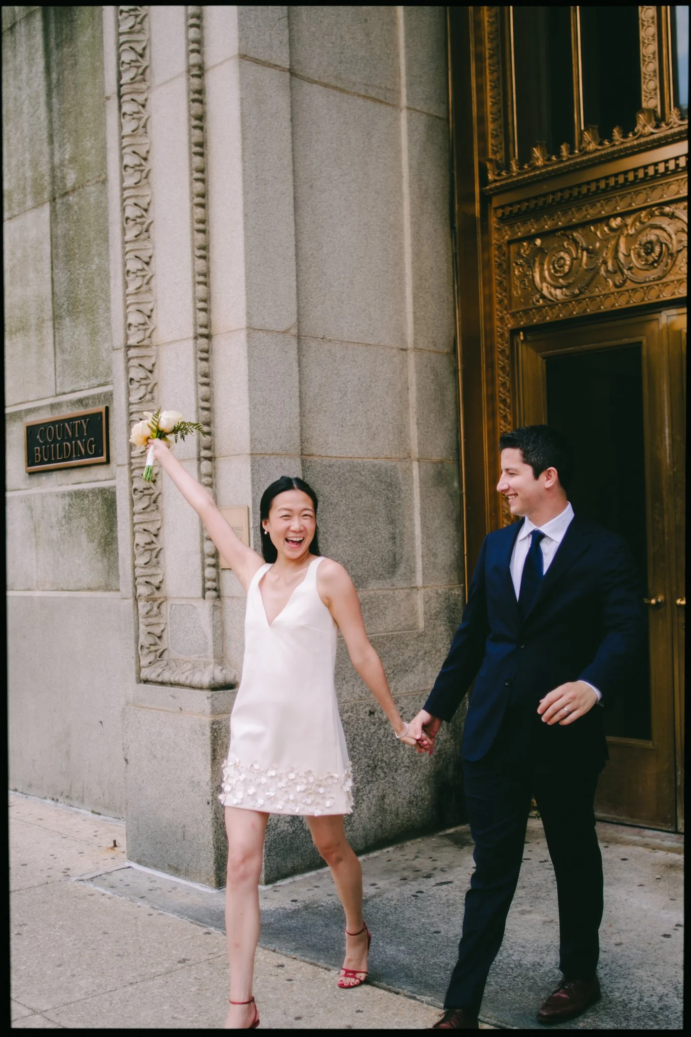 A woman in a white dress holding a bouquet and smiling, holding hands with a man in a navy suit and tie, both outside a building with ornate gold door and a sign reading 'County Building'.