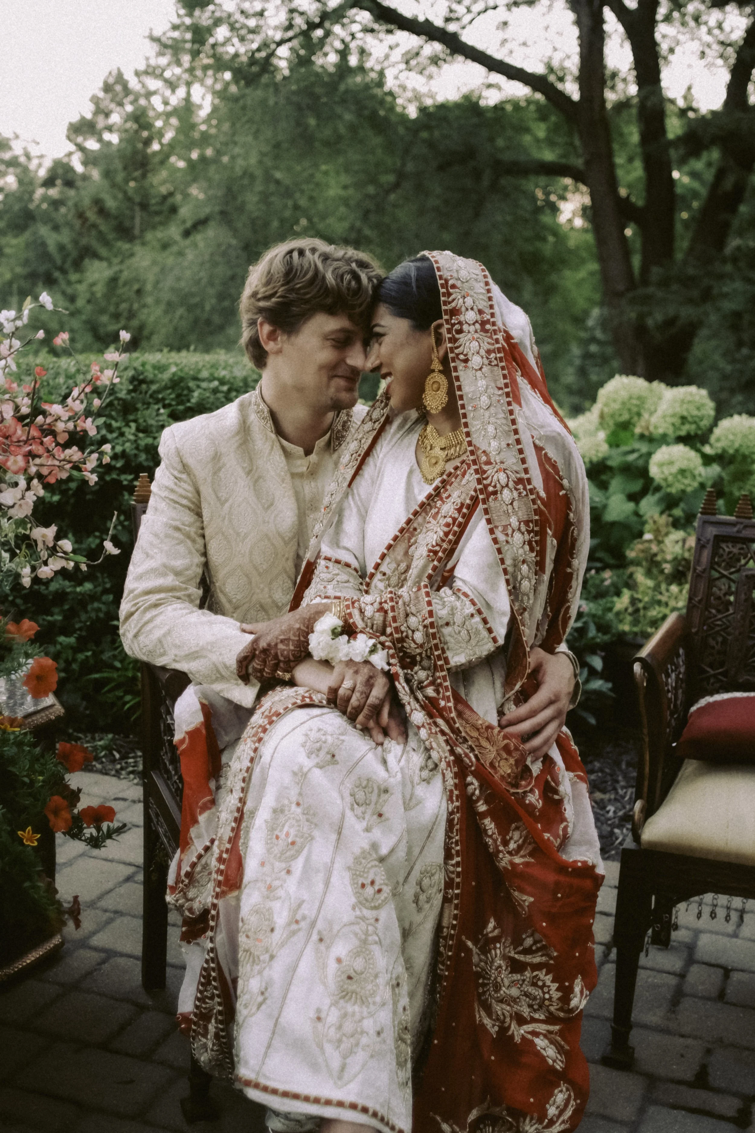 A South Asian bride and a Caucasian groom sharing a moment on their wedding day, seated outdoors among greenery and flowers, dressed in traditional wedding attire.