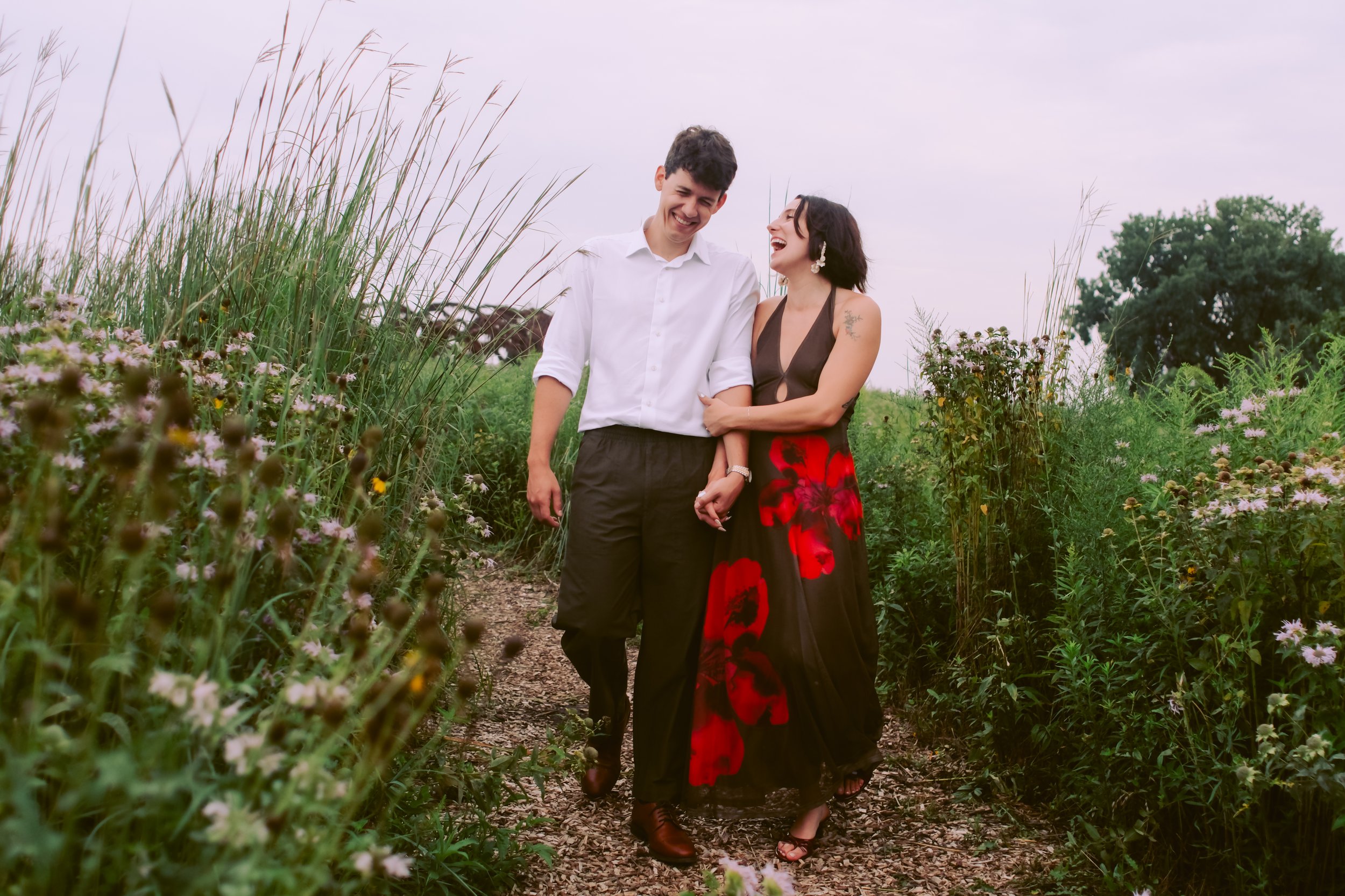 A man and woman walking on a dirt path through a field of tall grass and wildflowers, smiling and laughing together.