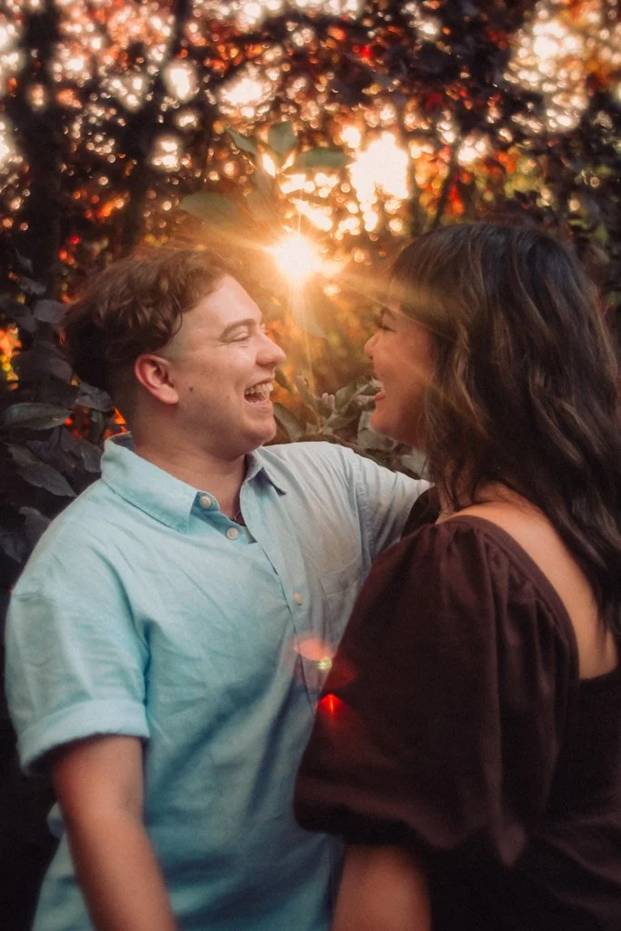 A happy couple smiling and gazing at each other outdoors during sunset, with sunlight filtering through trees in the background.