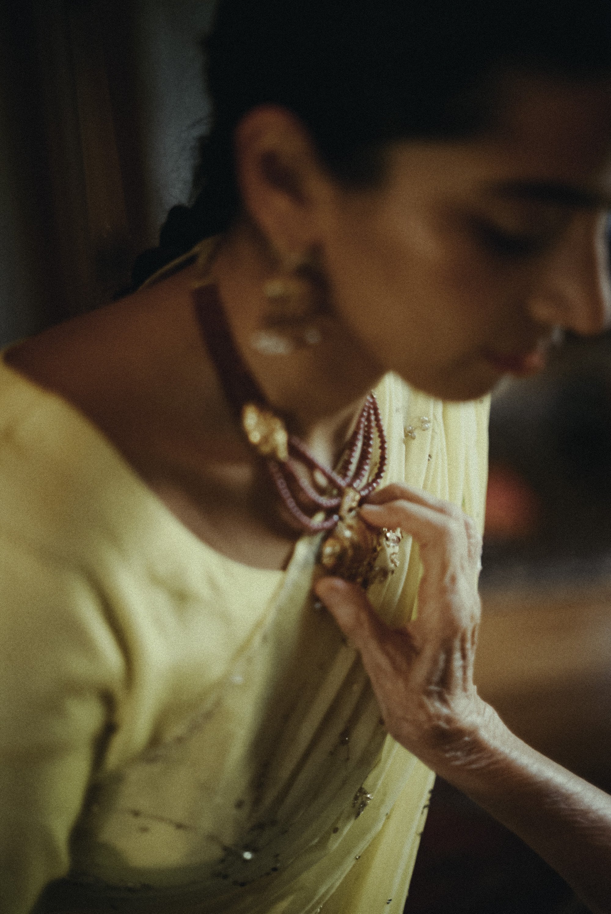 A woman dressed in yellow traditional attire wearing jewelry and being assisted by an elderly hand, possibly for prayer or a special occasion.