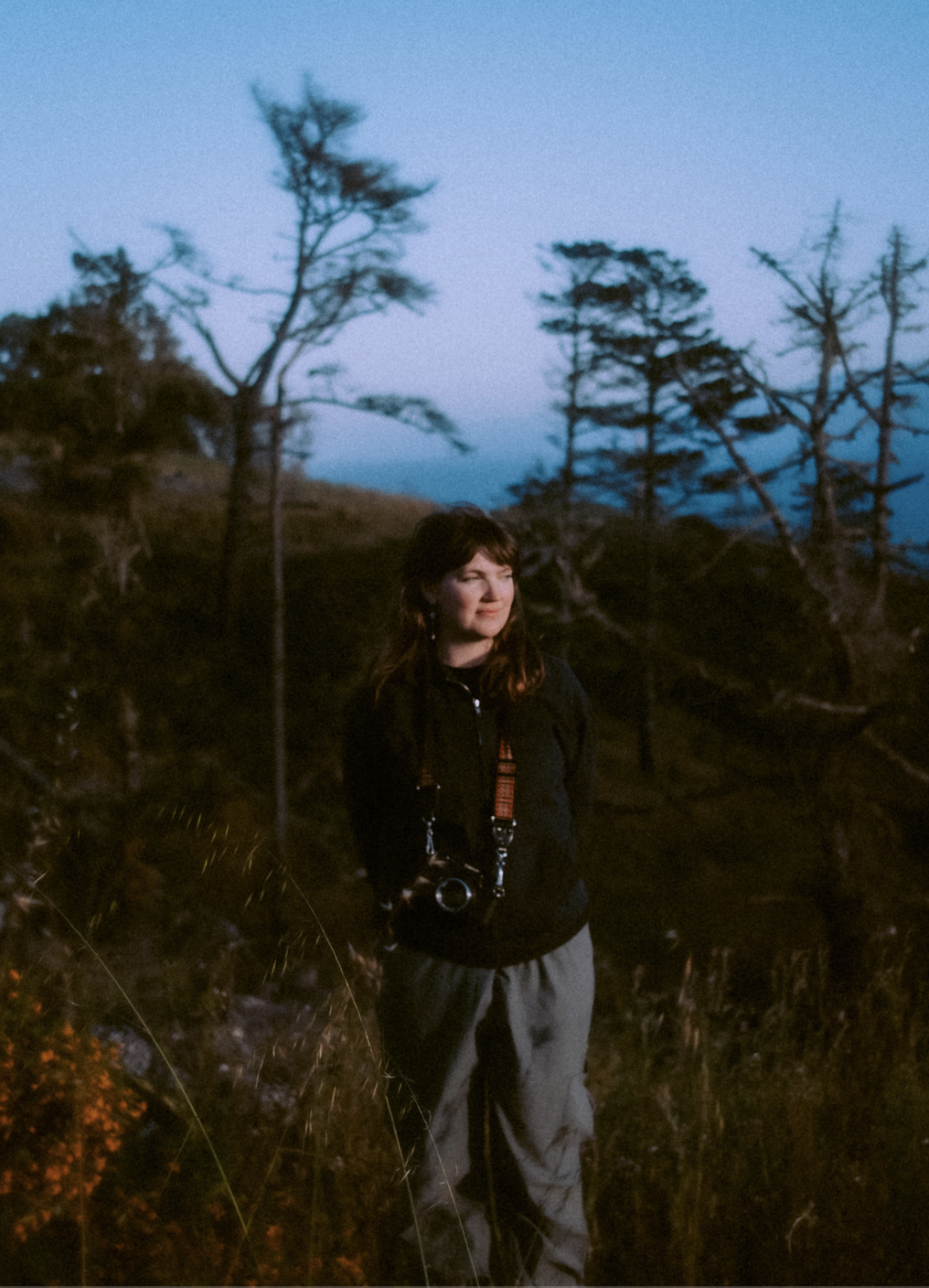 A woman standing outdoors in nature during dusk, surrounded by trees, wearing a black jacket and gray pants, with a camera hanging around her neck.