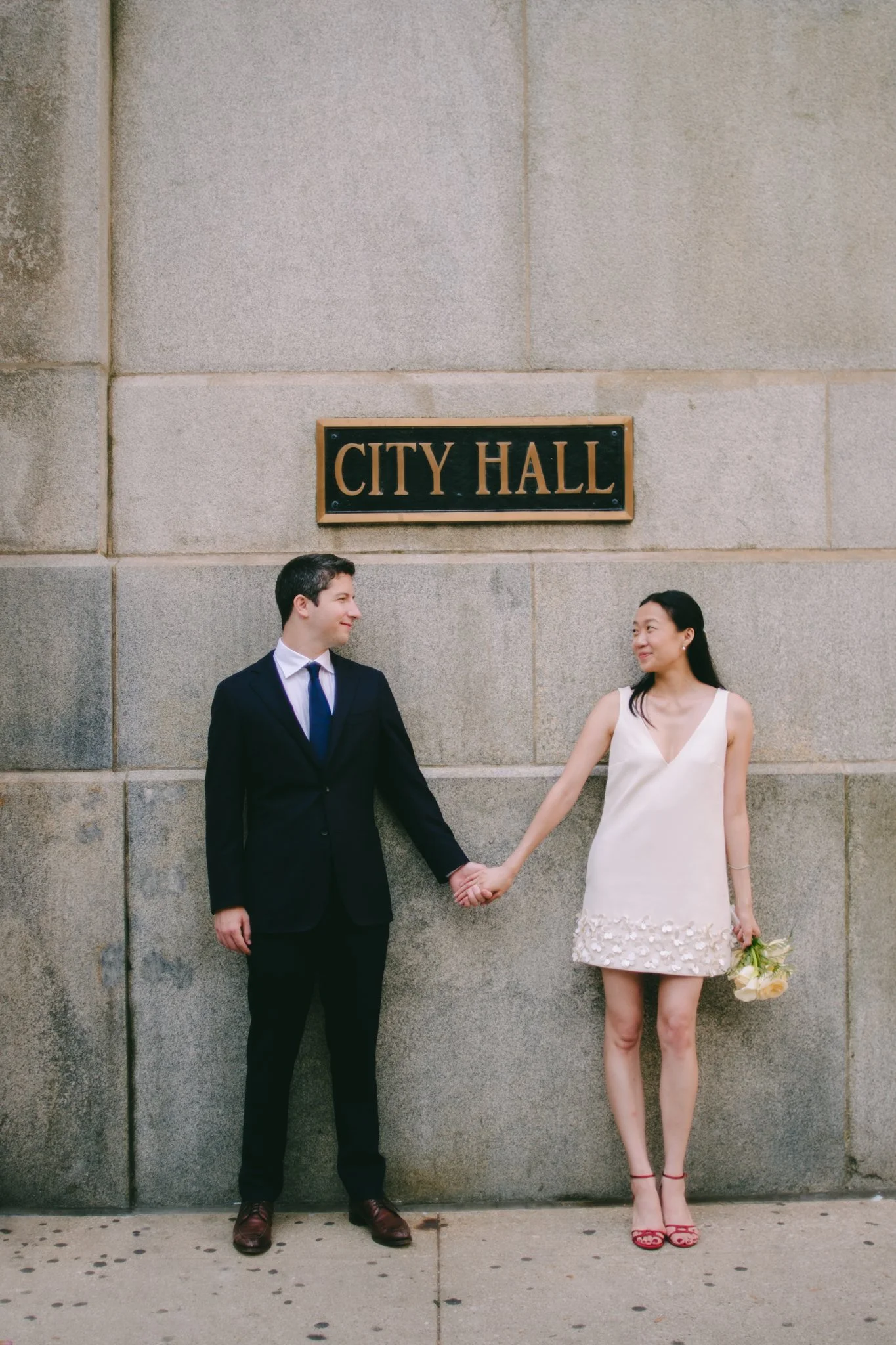 A man in a black suit and a woman in a white dress holding hands in front of a stone wall with a sign that says "City Hall". The woman is holding a bouquet of flowers.