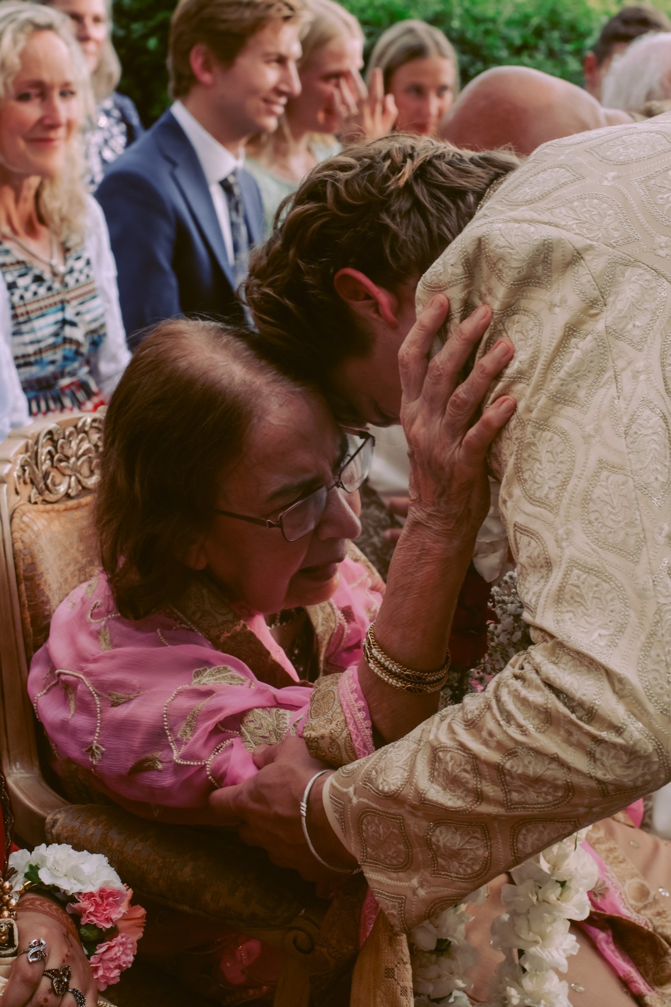 A woman in pink traditional attire and glasses is emotional with her eyes closed and tears, while a man in a cream embroidered outfit leans over her, touching her head during a ceremony, with other people watching in the background.