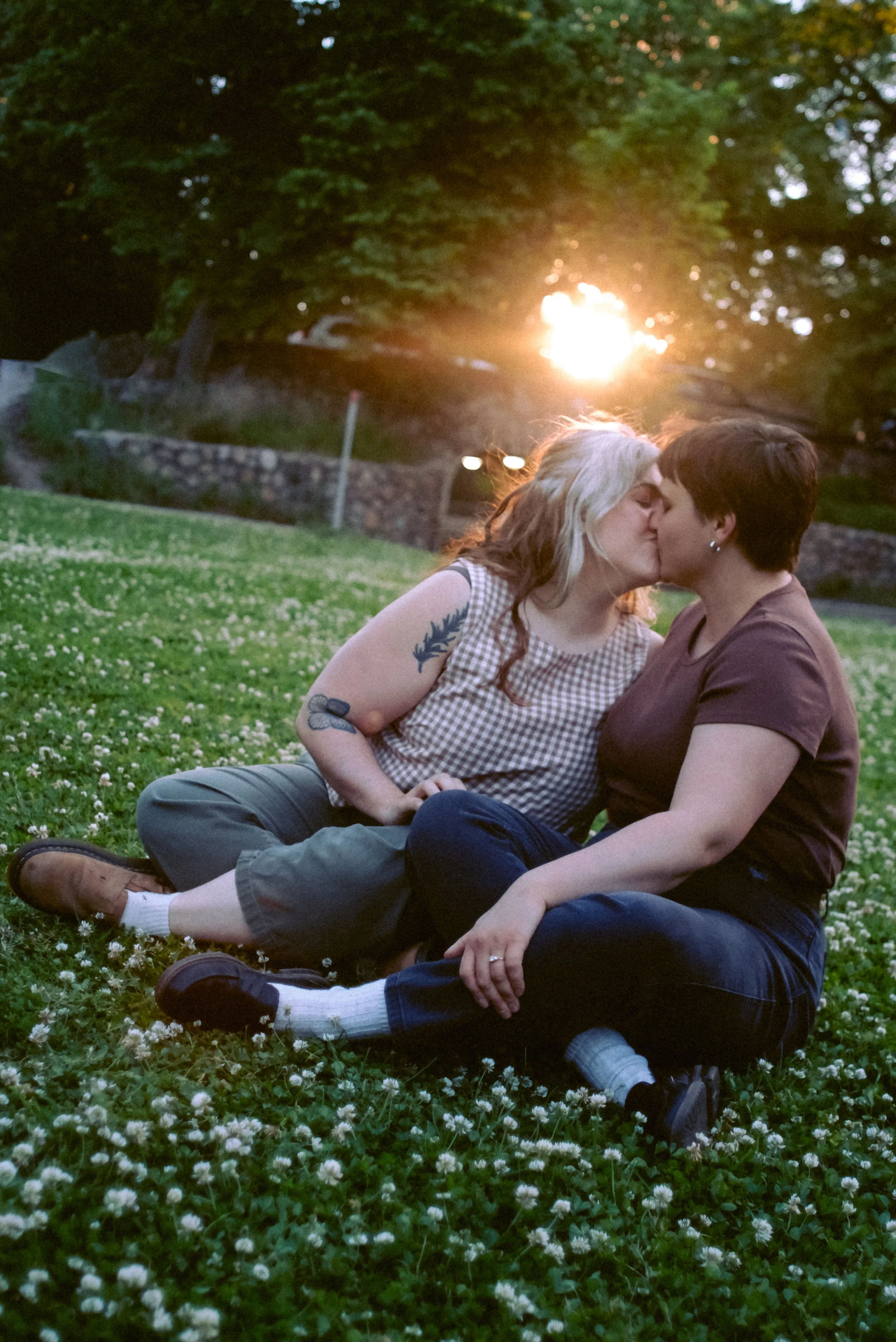 Two women sitting on grass with white flowers, kissing outdoors during sunset, surrounded by trees.