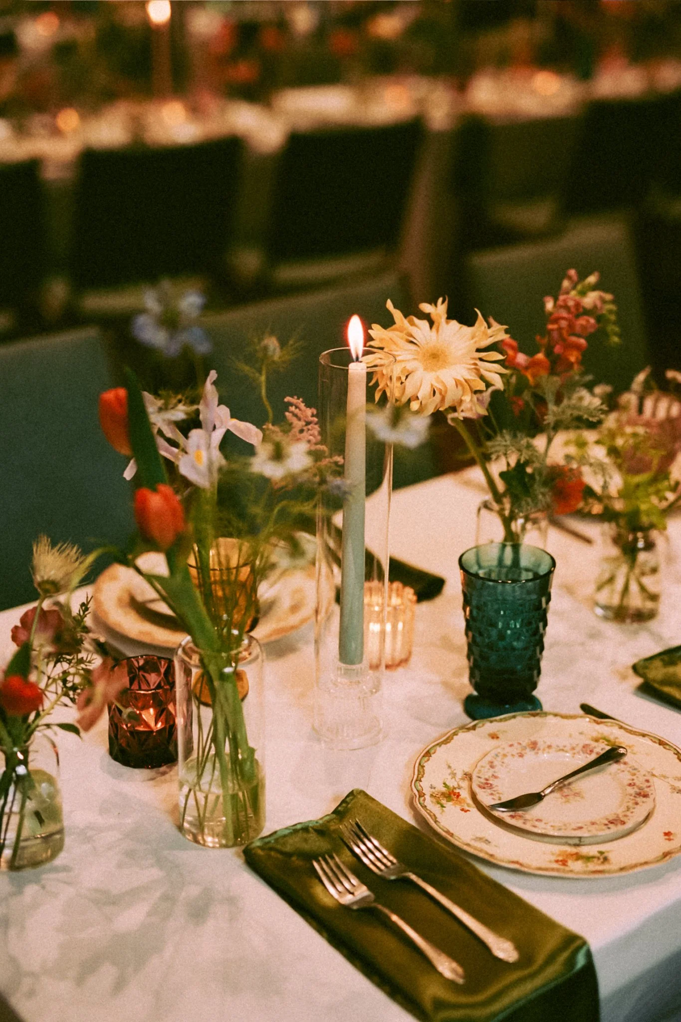 A festive table setting with floral arrangements, a lit candle, decorative plates, and cutlery on a white tablecloth, with a blurred background of chairs and warm lighting.