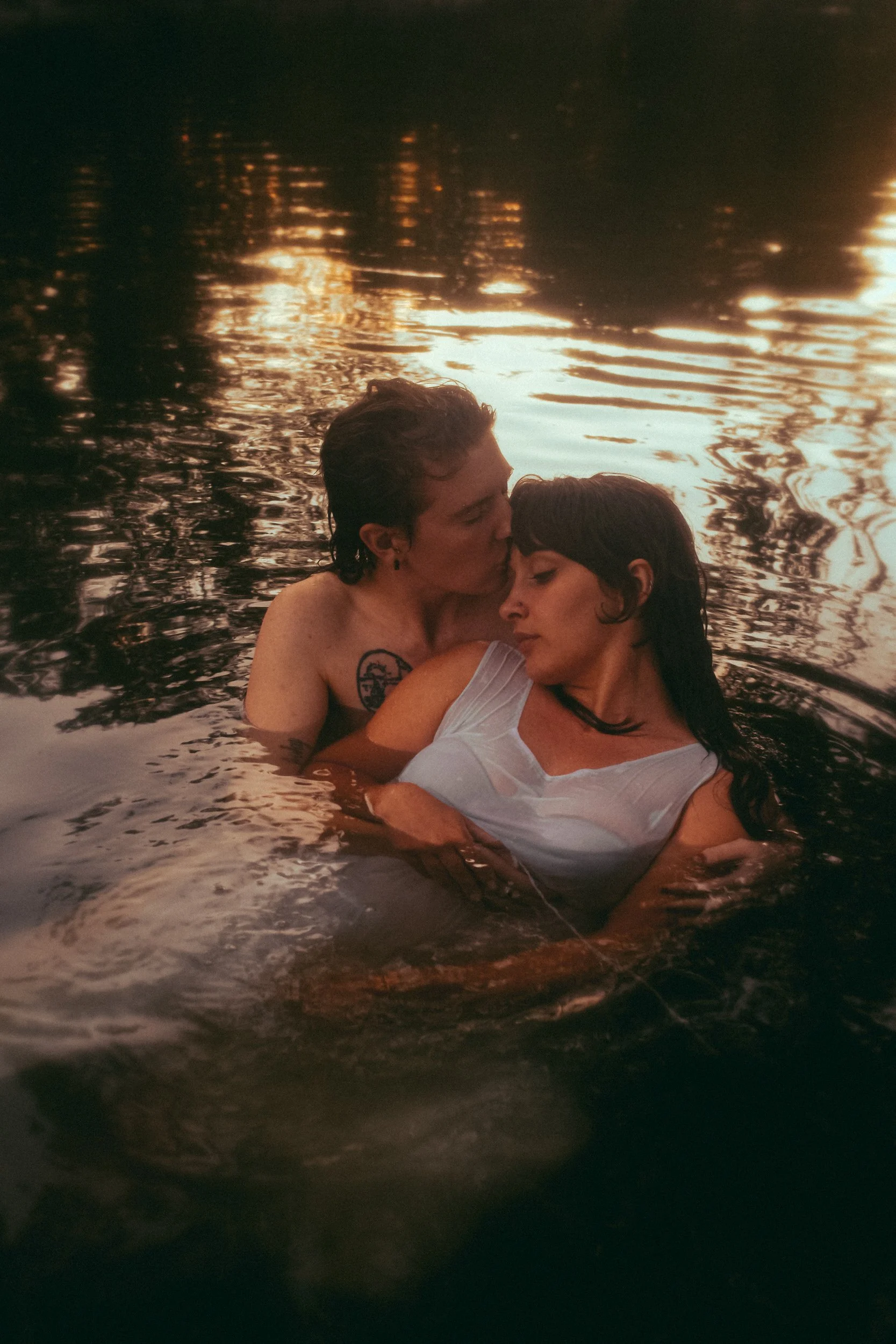 Two women are embracing in a body of water during sunset, with one woman wearing a white top and the other with short hair and tattoos.