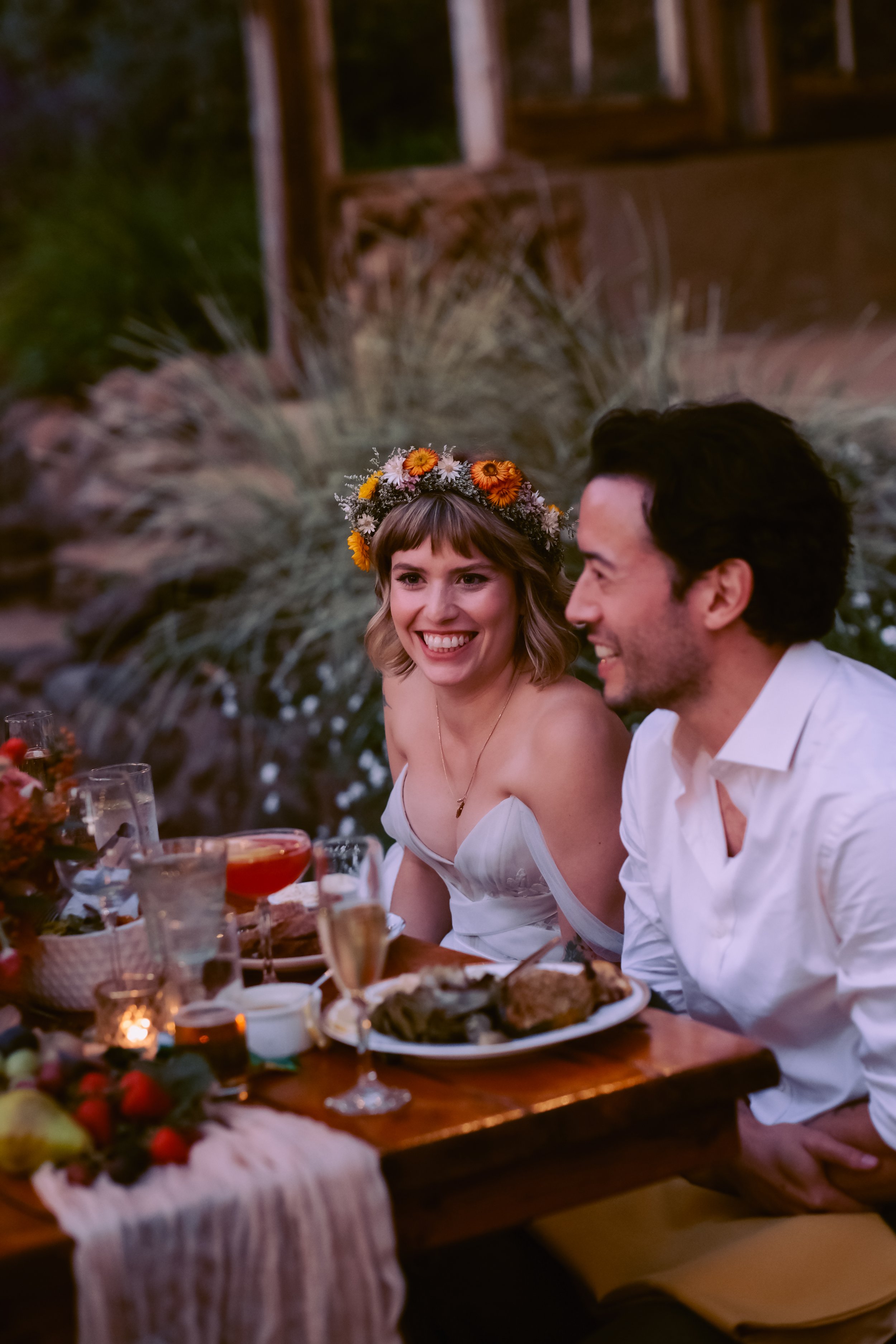 A woman with a flower crown smiling next to a man at a dinner table outdoors during evening, with various foods and drinks, in a decorated setting surrounded by plants.