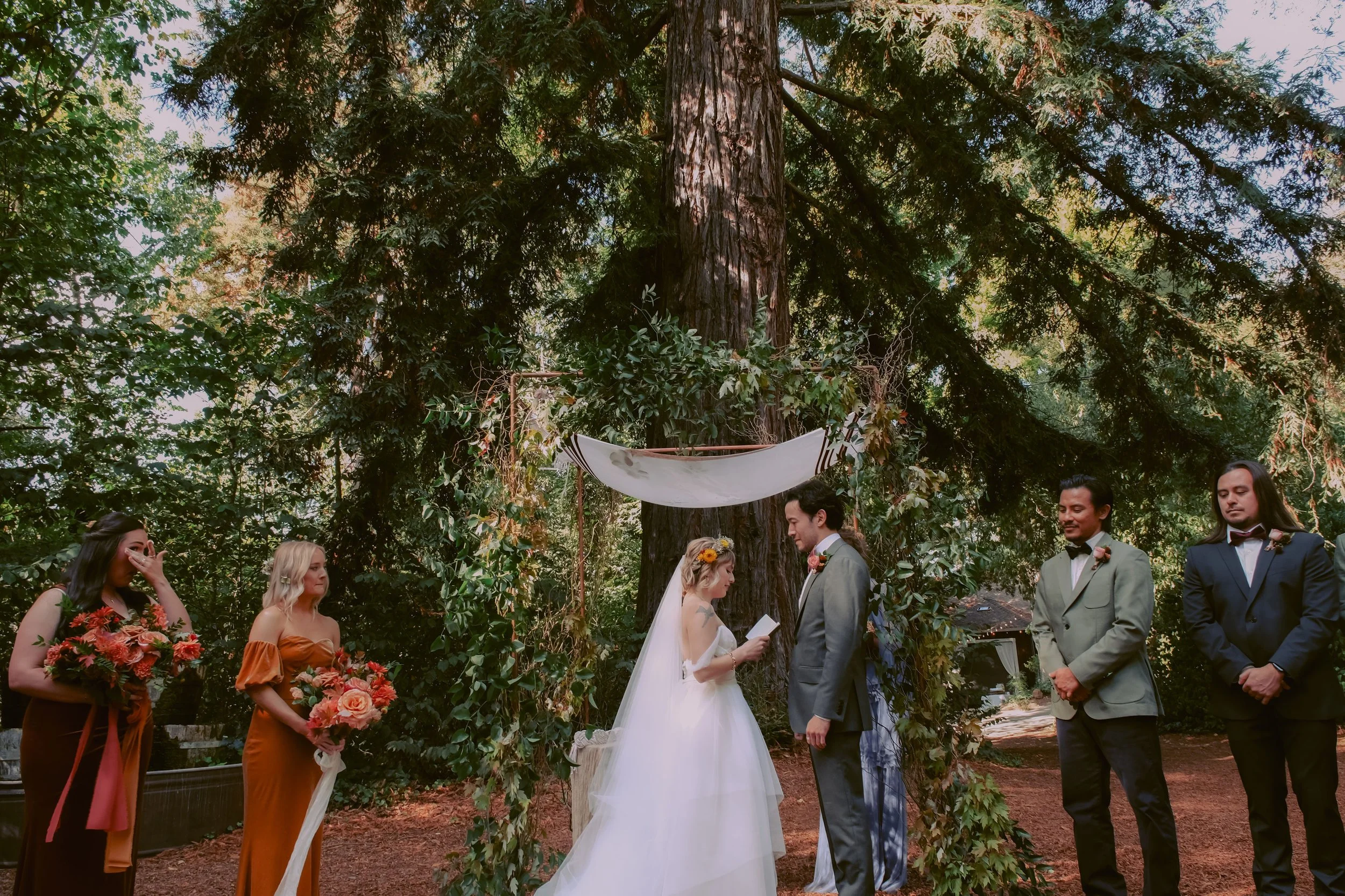 A wedding ceremony outdoors beneath a large tree, with six people present. The bride, in a white gown with a veil and flower crown, reads vows to the groom, who is in a gray suit. Two bridesmaids hold bouquets of pink and red flowers, and two groomsm