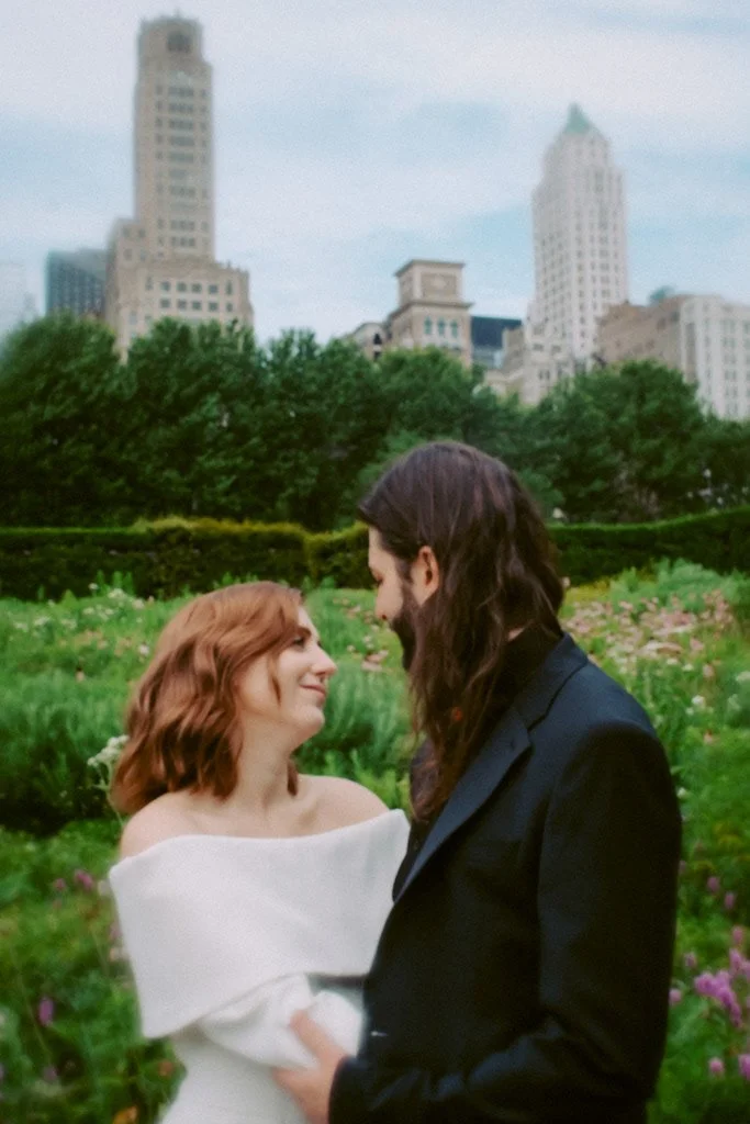 A couple with long hair sharing a romantic gaze in a lush garden with city buildings in the background.
