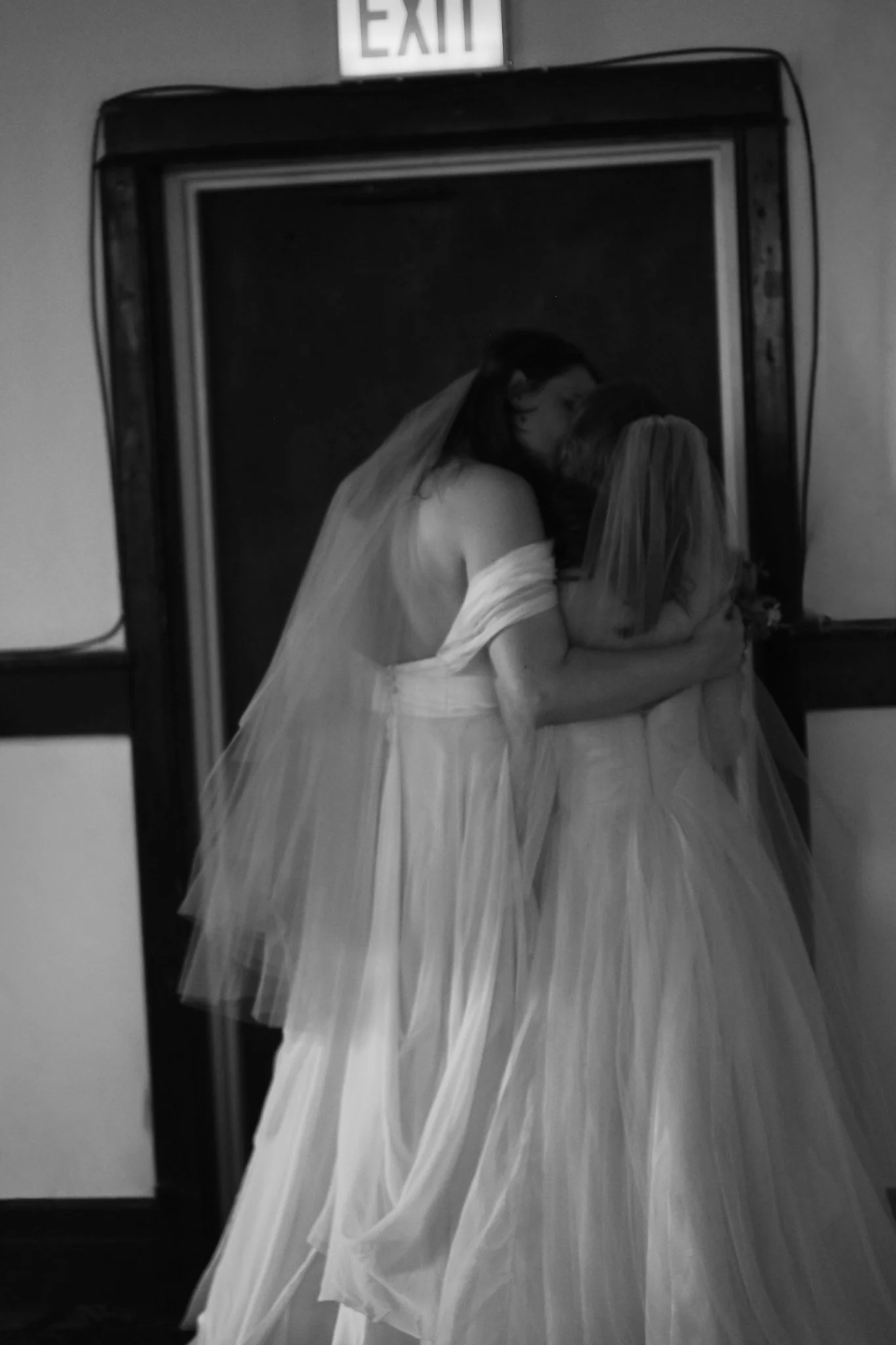 A black-and-white wedding photograph of a bride and groom sharing a kiss, reflected in a mirror with an exit sign above.