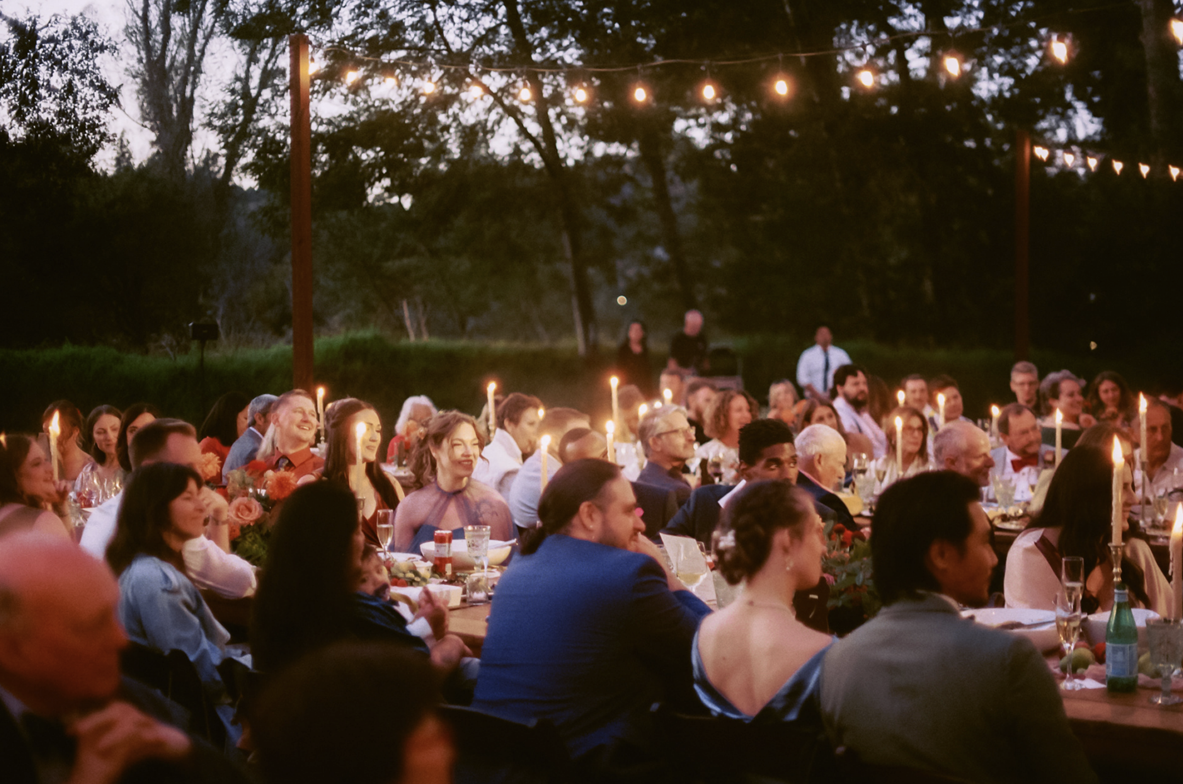 People sitting at outdoor dinner table during evening with string lights overhead and candles on the table, enjoying a celebration in a natural setting.