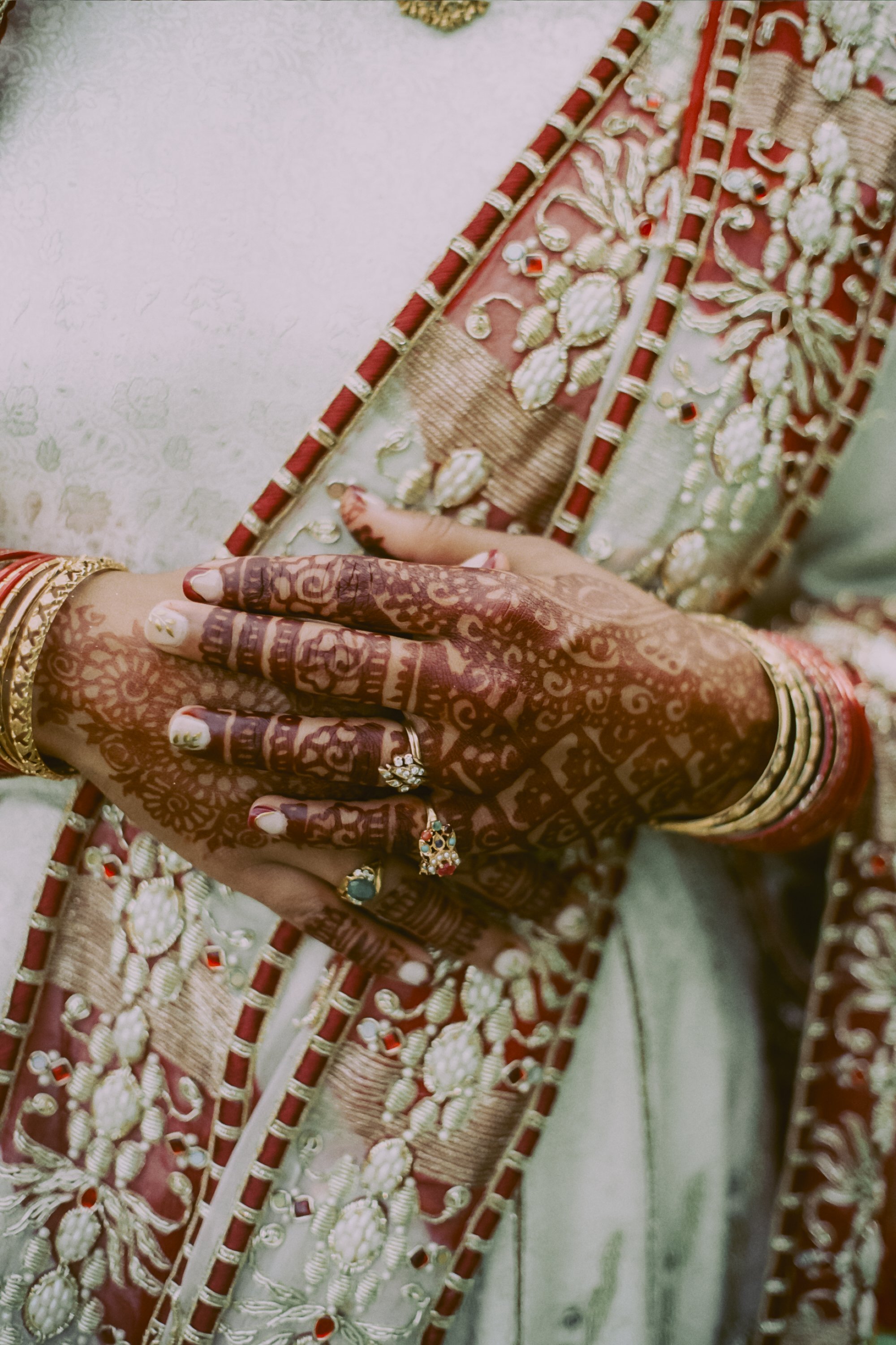 Close-up of hands adorned with henna, rings, bangles, and gold jewelry, held together in a prayerful pose against a richly embroidered saree.