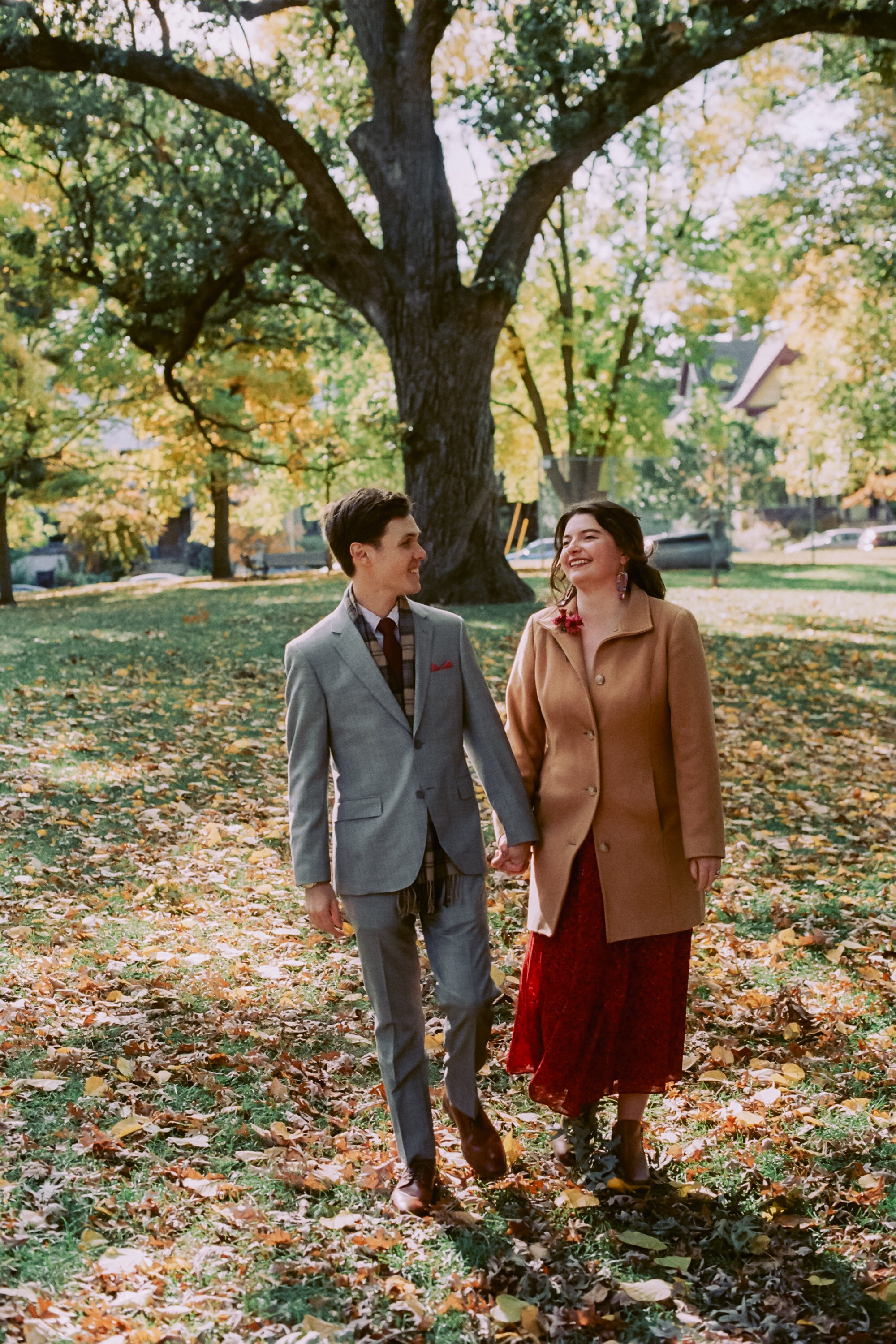 A young man and woman walk hand in hand through a park covered in fallen leaves during autumn, smiling and looking at each other with tall trees and houses in the background.