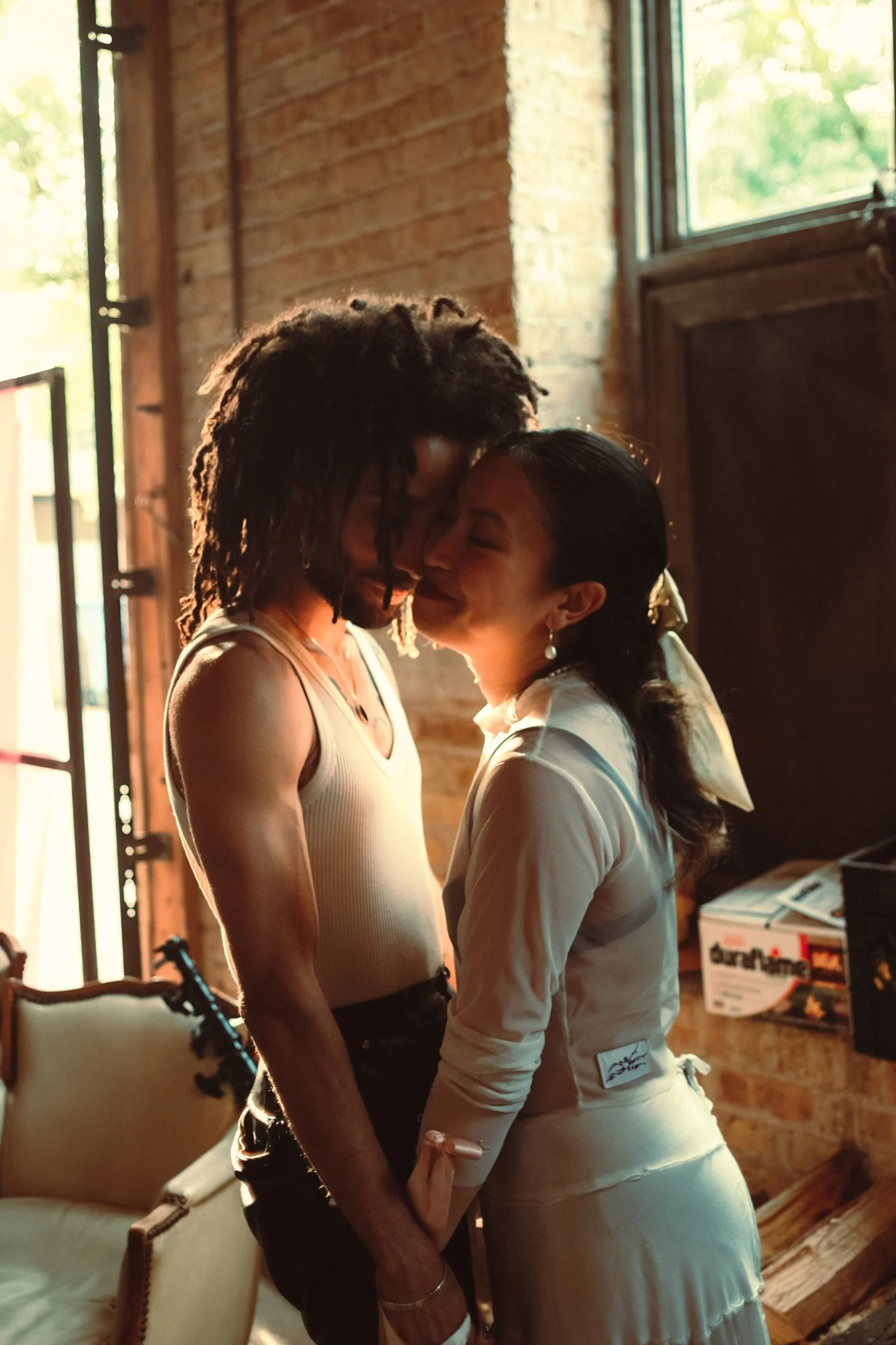 A couple standing close together indoors, touching foreheads and smiling with eyes closed, warm natural lighting.