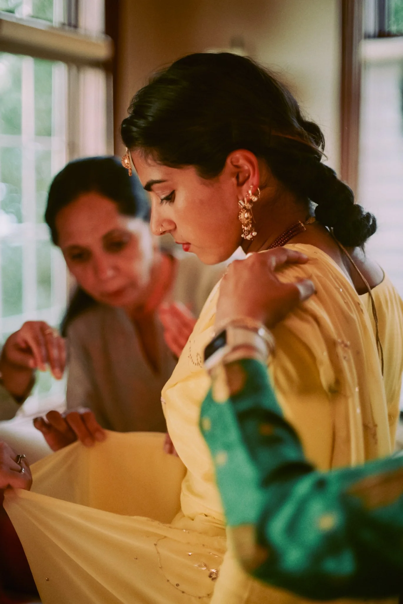 Two women in traditional Indian attire, one adjusting her yellow dress, with ornate jewelry, in a setting with natural light coming through windows.