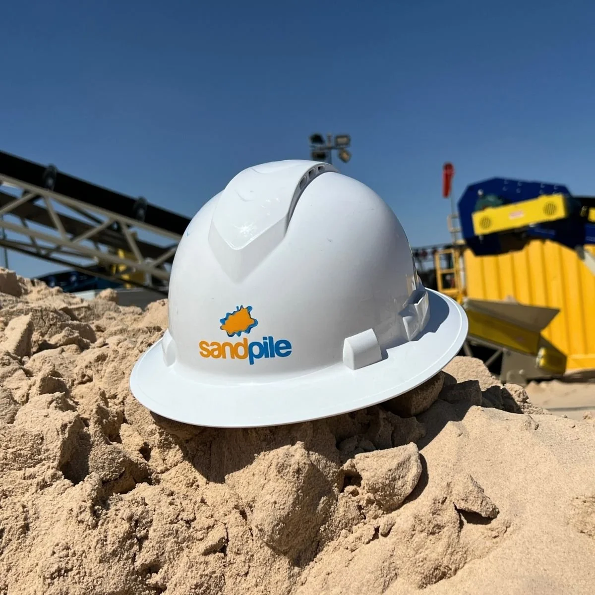 white construction helmet with 'sandpile' logo resting on sand at a construction site with machinery and equipment in the background in Midland, Tx.