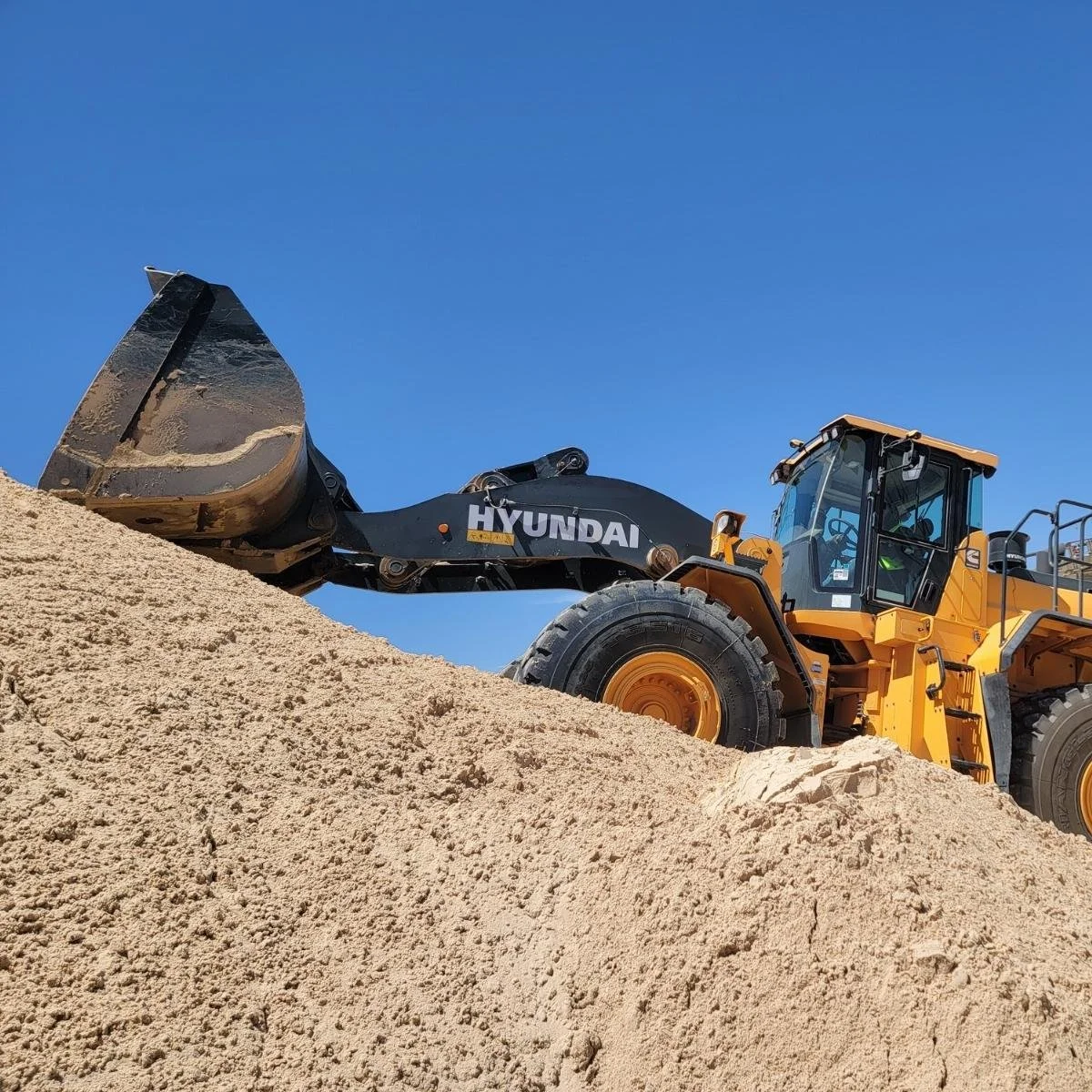 A yellow Hyundai front loader is scooping sand at a construction site under a clear blue sky in Midland, TX.