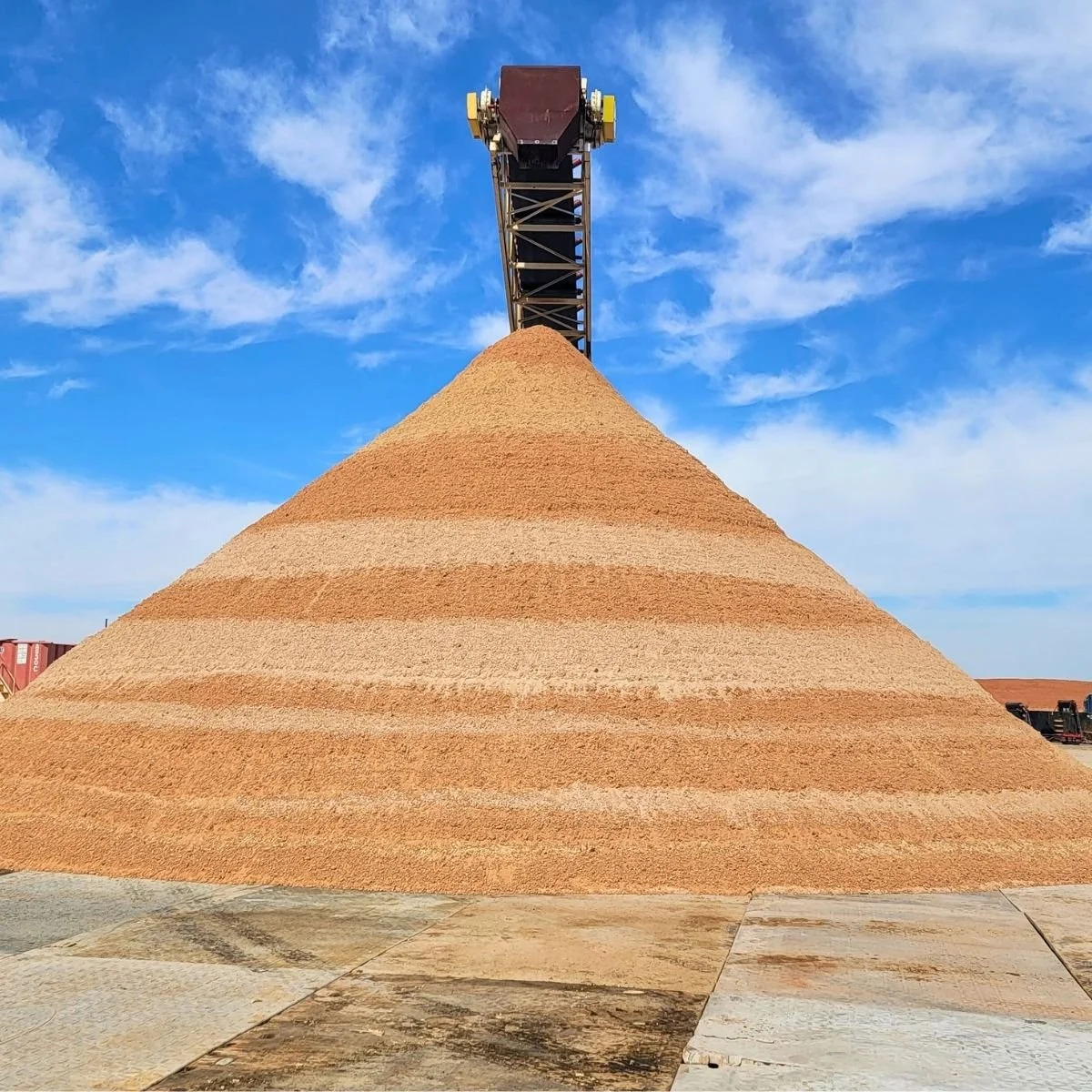 A large conical pile of sand in the Permian Basin with a machinery crane featuring a scoop at the top behind it, set against a blue sky with scattered clouds in Midland, Texas.