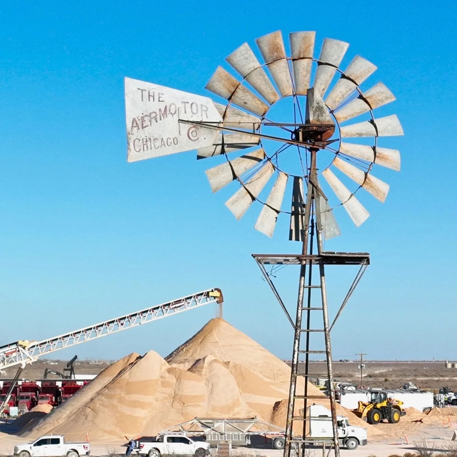 An old windmill with the sign 'The Aeromotor Chicago' in a desert landscape with piles of sand and construction equipment in Midland, Tx and Permian Basin.