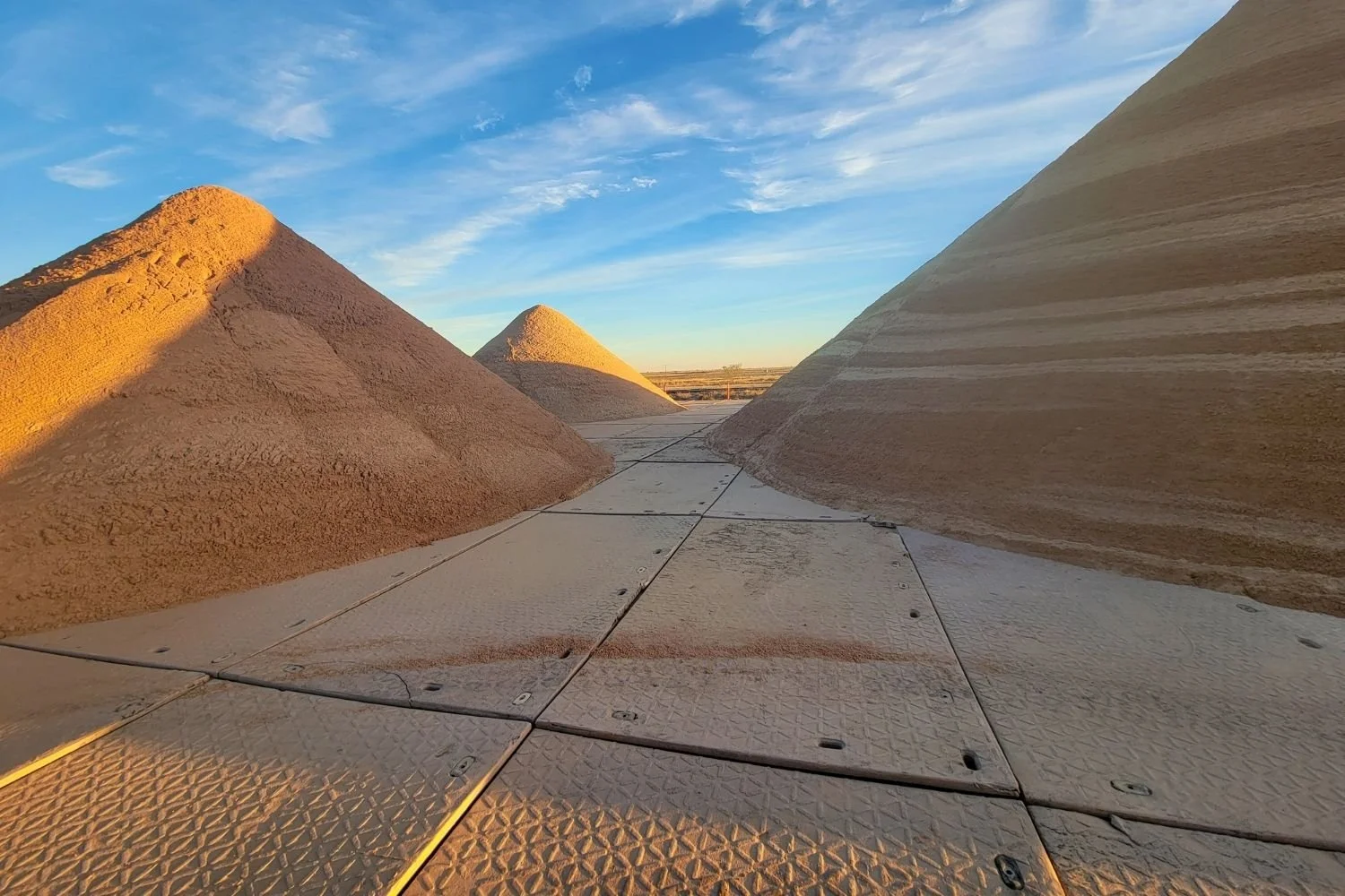 Large piles of pink and beige salt stacked in a desert landscape under a blue sky with clouds, with a patterned metal surface in the foreground.