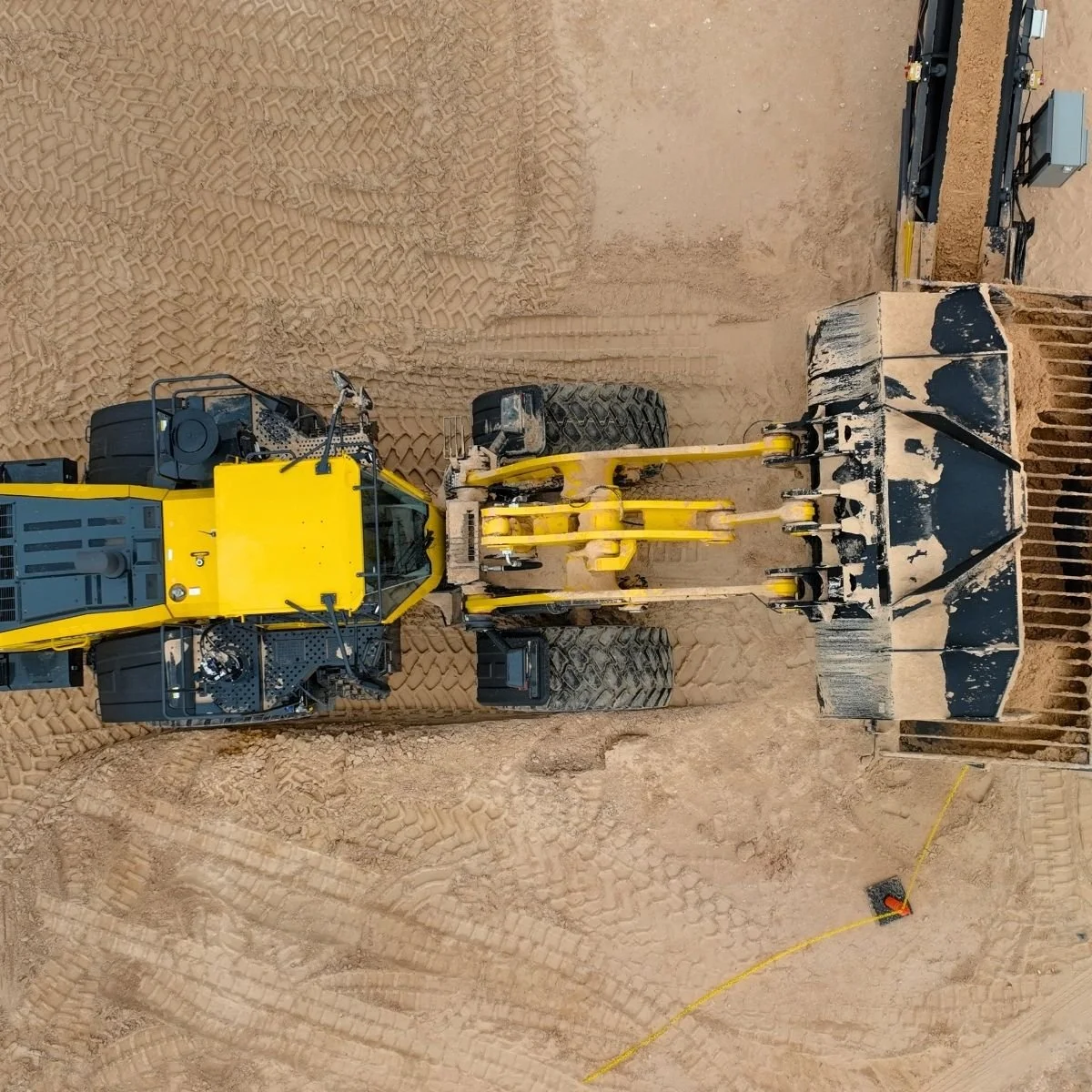 A large yellow construction vehicle on a SandPile location in Eagle Ford, with a wide black and yellow scoop at the front, moving frac sand and leaving tire tracks in the soil near Midland, Texas.