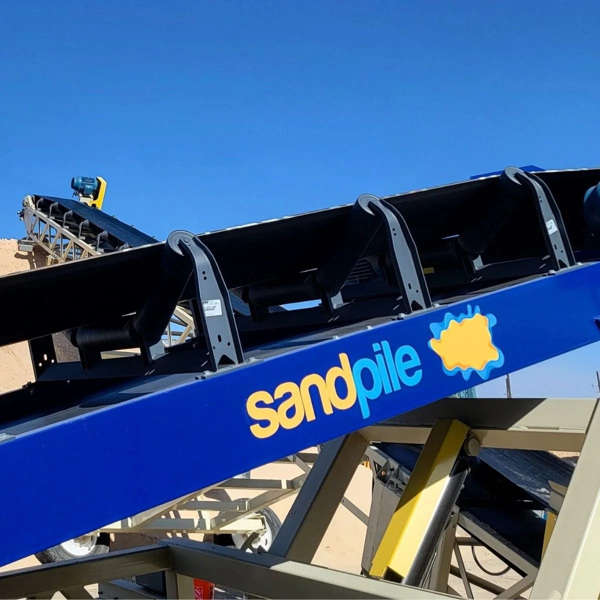Close-up of a sandpile conveyor belt with bright blue background, showing black rotating discs and a blue structure labeled "sandpile" with a sand in Midland, TX.