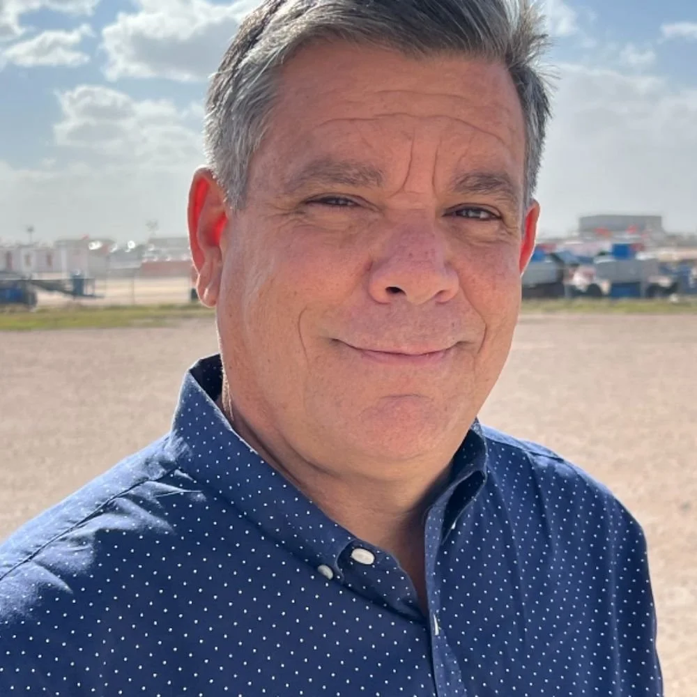 A man with short, gray hair smiling outdoors on a sunny day, wearing a blue polka dot shirt, with a blurred cityscape and parked cars in the background in Midland, Texas.
