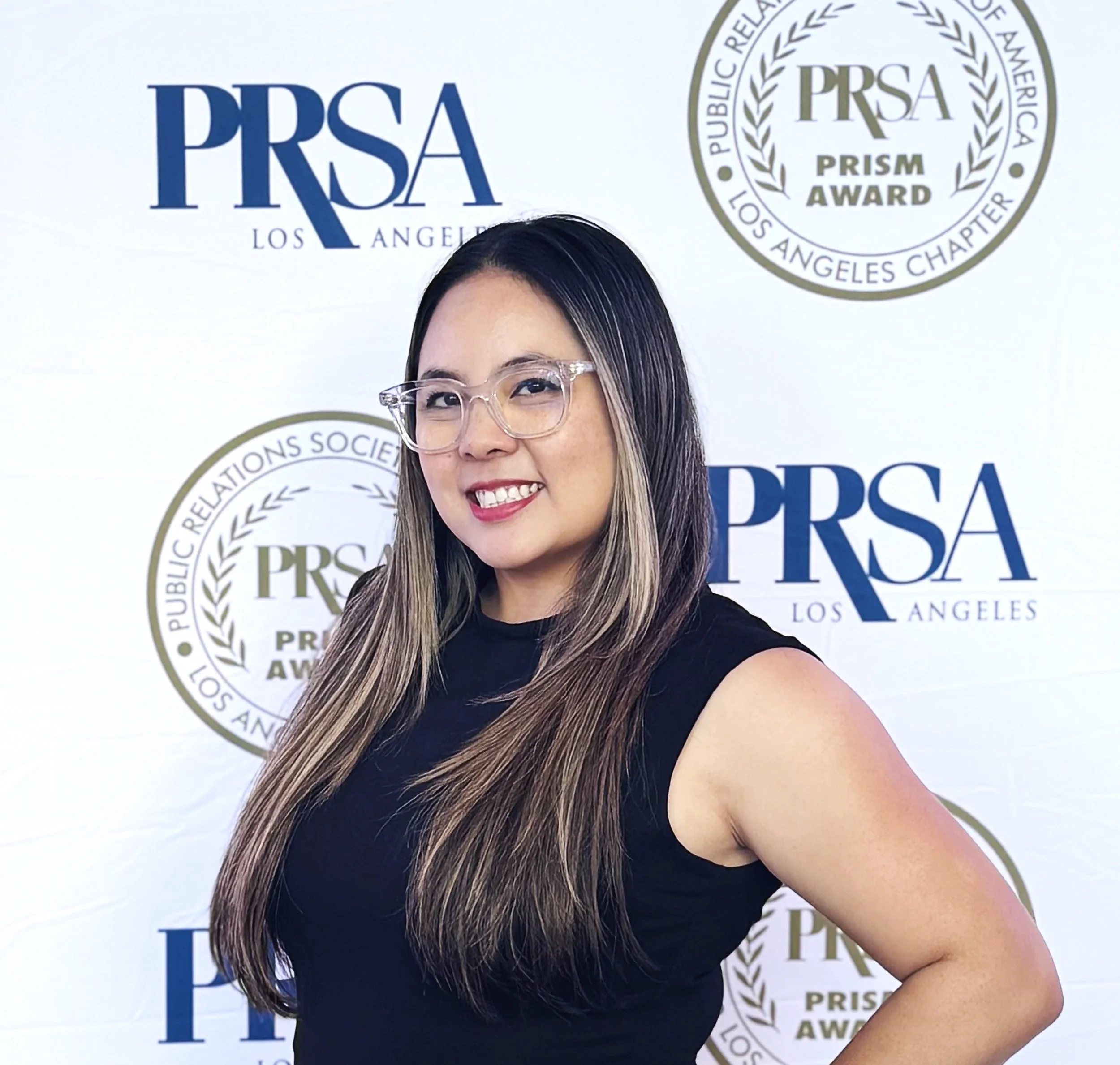 A woman with long, straight hair and glasses, smiling at the camera, standing in front of a backdrop with the PRSA Los Angeles logo and PRISM Award branding.
