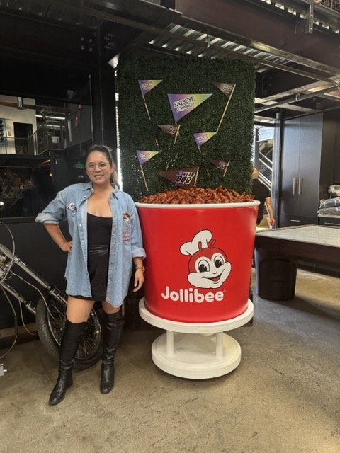 Ashley, founder, stands next to a giant Jollibee fried chicken bucket display at the FOODBEAST x Jollibee Drippin' Event, with a green backdrop and decorative flags.