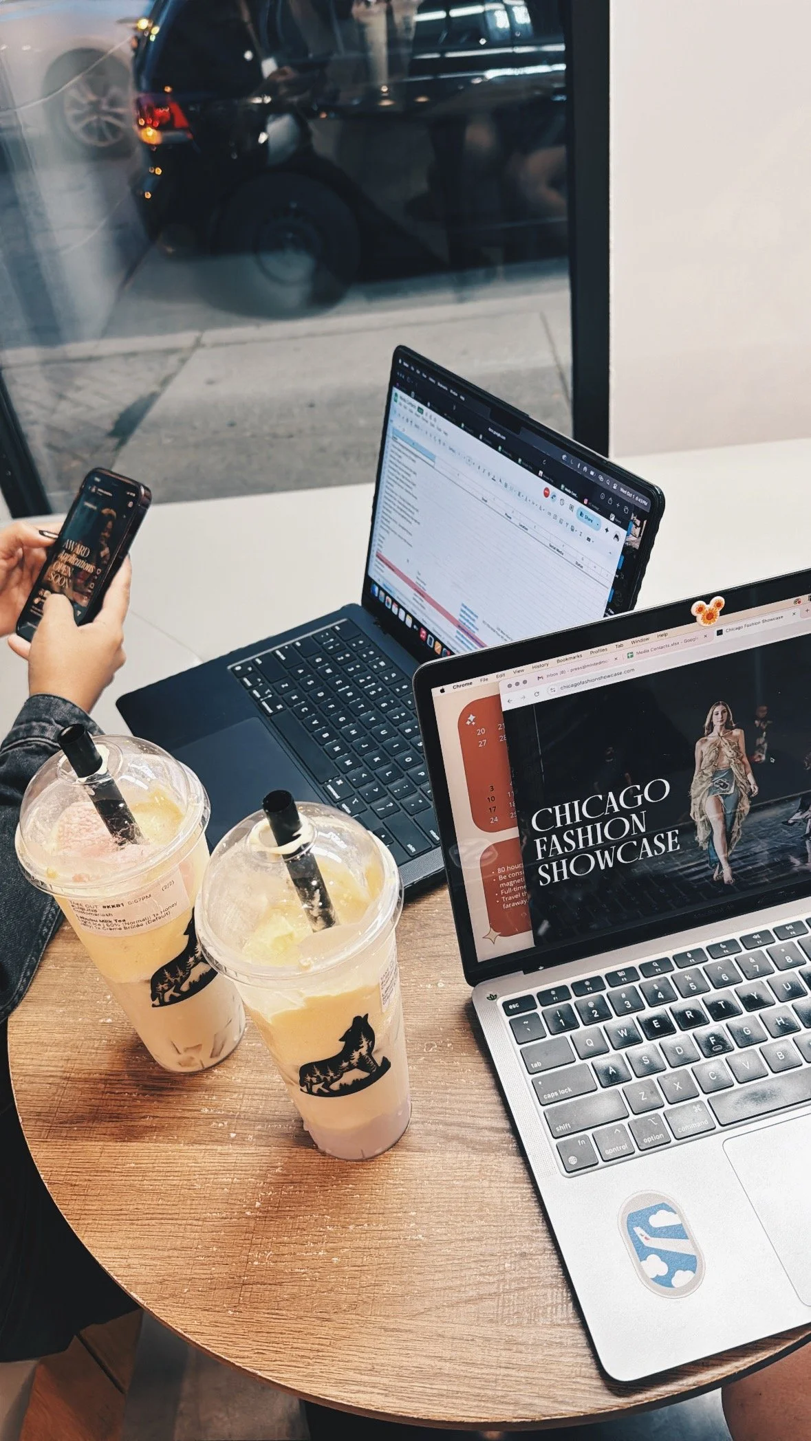 A person working on a laptop and phone at a table with two drinks in Chicago, IL near a window showing parked cars outside. The two members are discussing the Chicago Fashion Showcase.