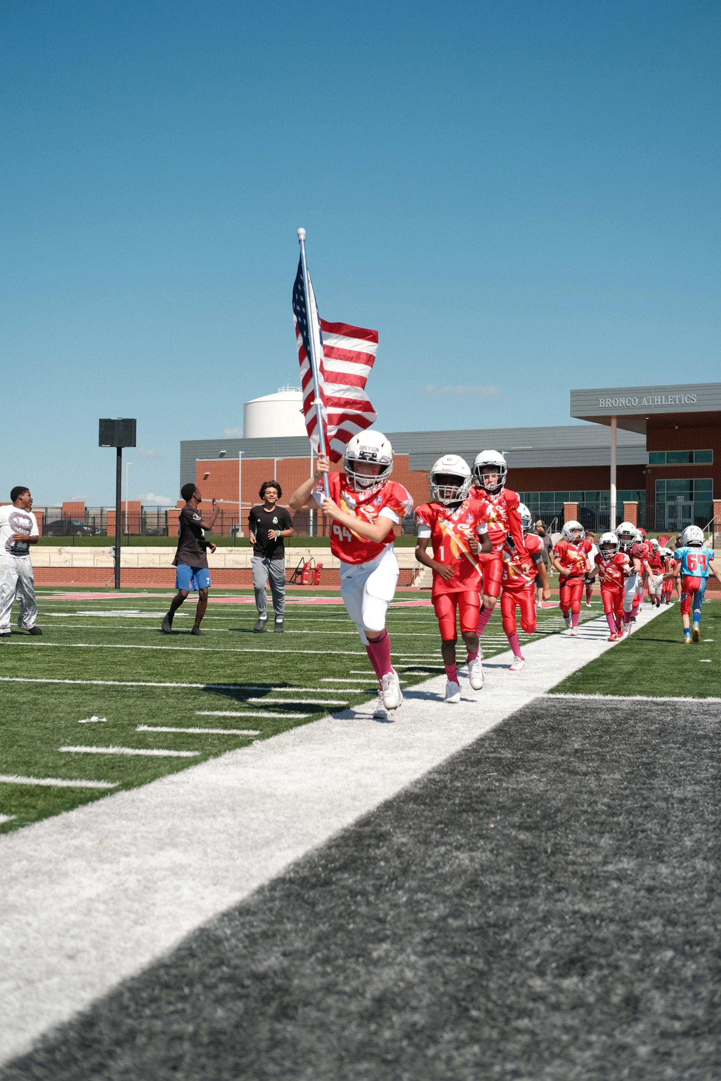 Group of young football players in red uniforms running onto the field while the player at the front carries an American flag, with a clear blue sky and school building in the background.