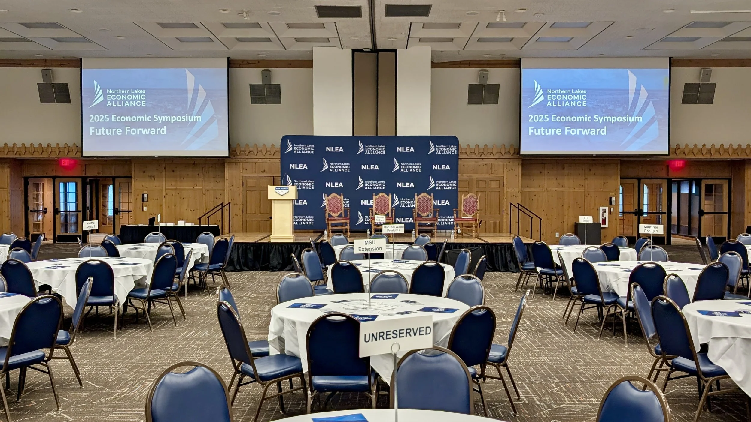 Conference room set up for an event with round tables covered in white tablecloths and blue chairs. A stage with a podium and three chairs is at the front, with a backdrop displaying the Northern Lakes Economic Alliance logo. Two large screens above 