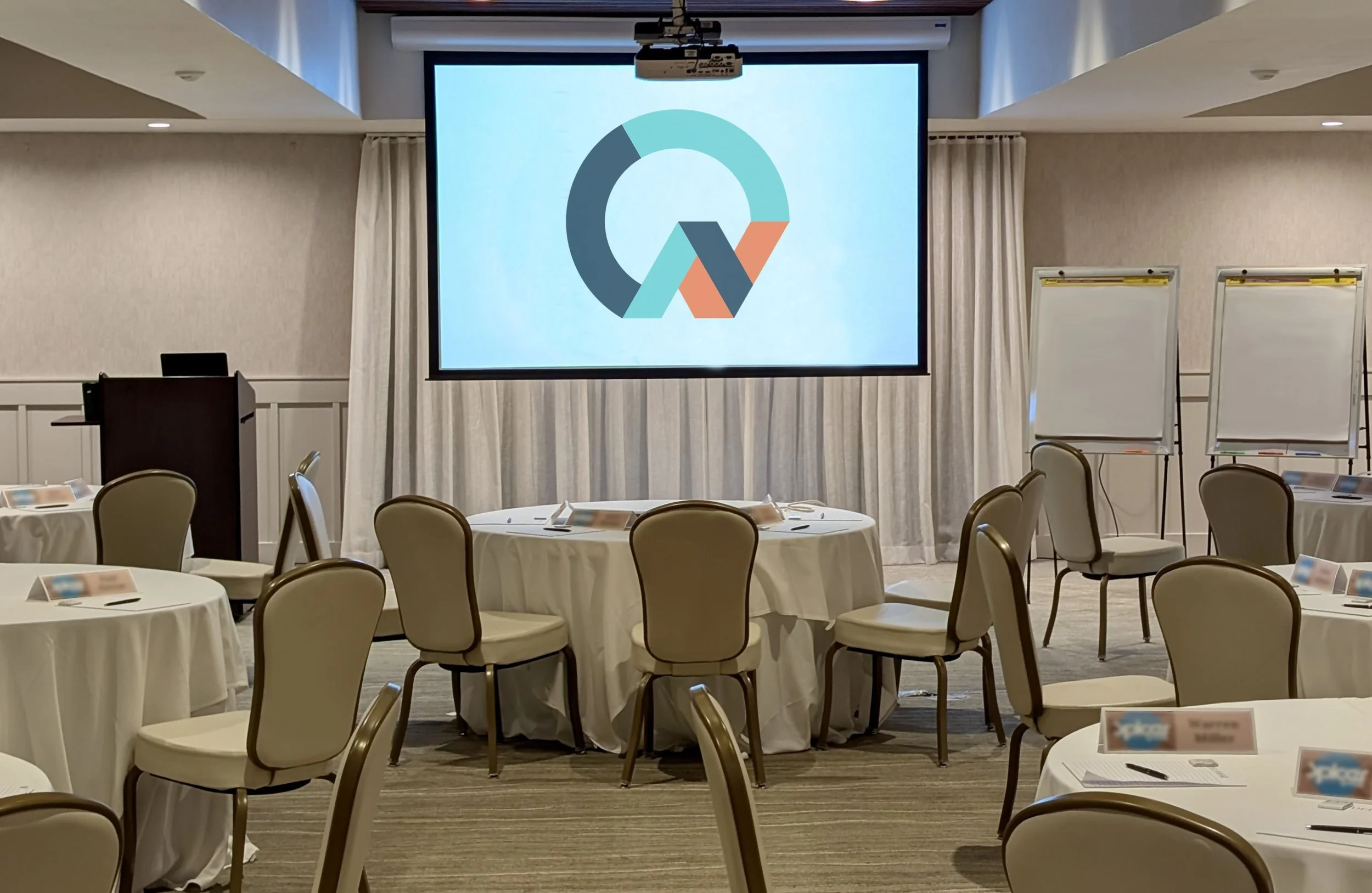 Conference room with round tables covered in white tablecloths and beige chairs, whiteboards on stands, and a large projection screen displaying a colorful logo, all in a well-lit room.